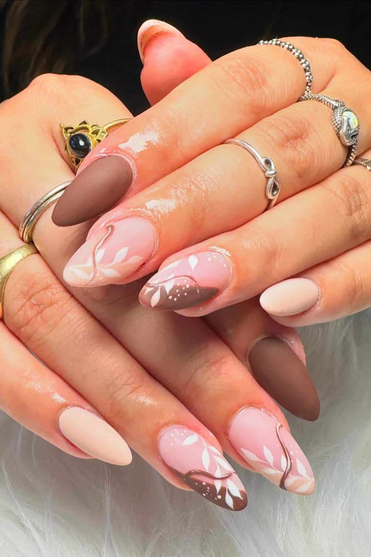 Hands with long almond-shaped nails featuring a pink and brown color scheme, abstract designs, and white leaf patterns. The person is wearing multiple rings.