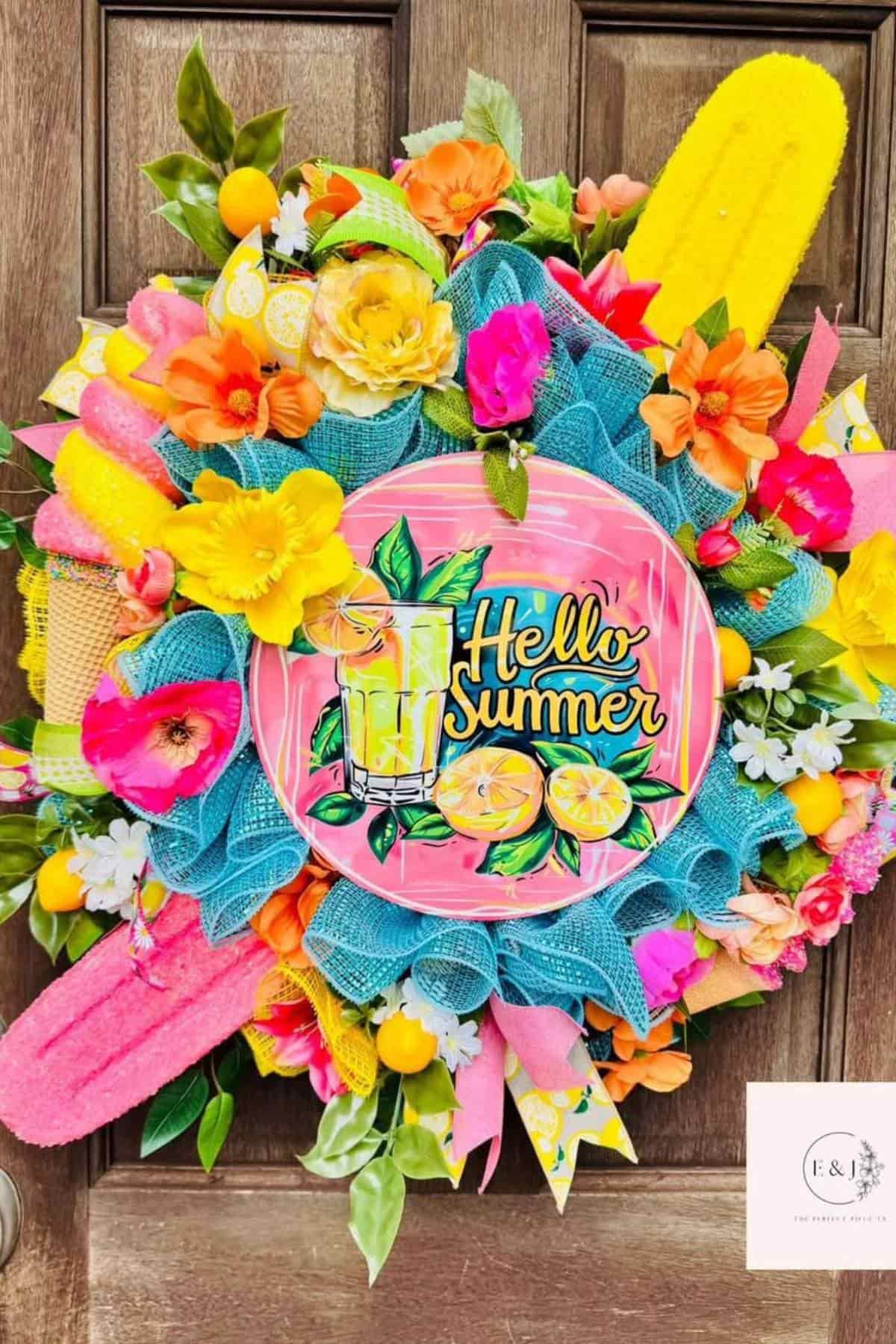 Colorful summer wreath with artificial flowers, lemons, popsicles, and a plate reading "Hello Summer" with a lemonade illustration, displayed on a wooden door.