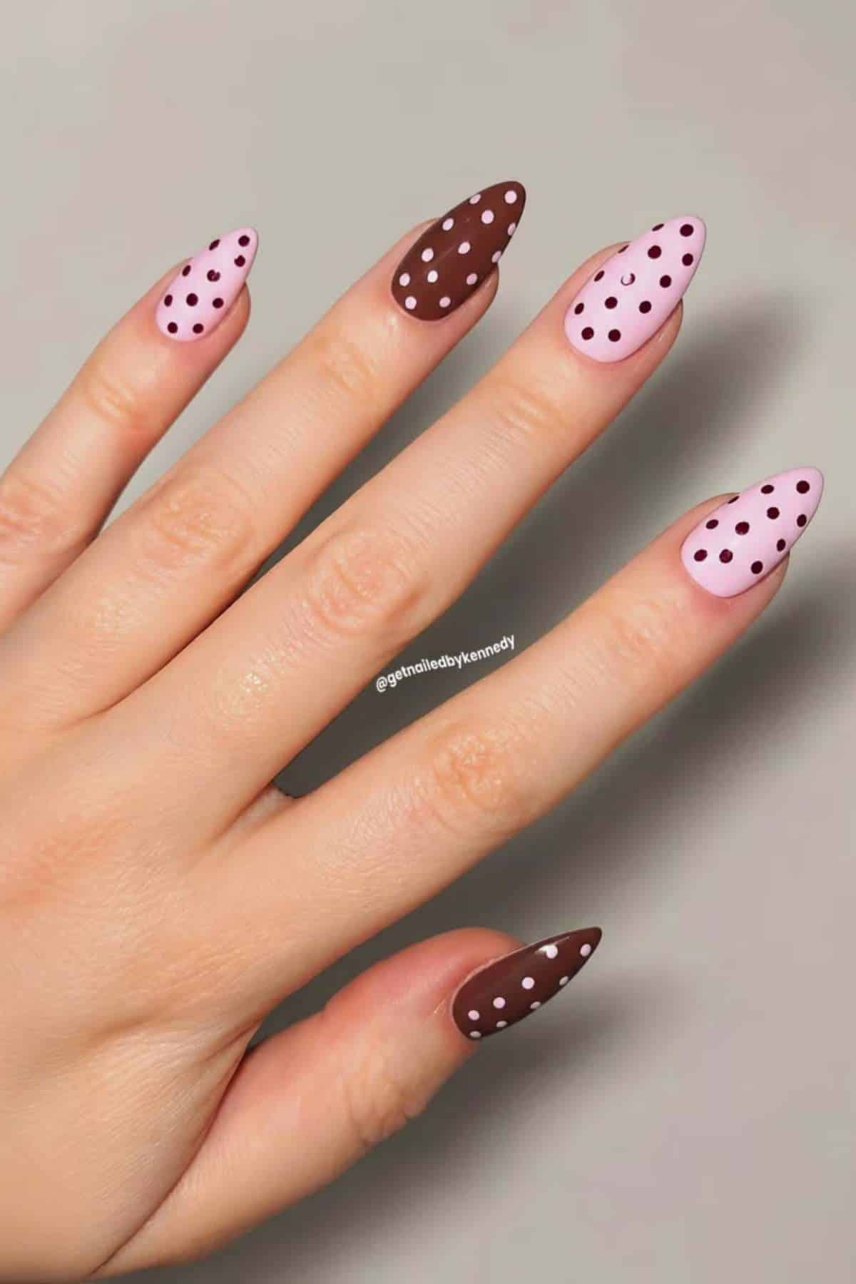 A hand with almond-shaped nails painted in alternating brown and pink colors, each nail decorated with polka dots of the opposite color.