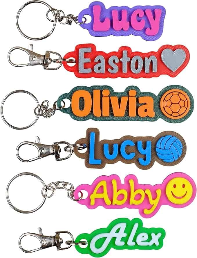 Six colorful name keychains are displayed, each with different names and designs: Lucy, Easton, Olivia, Lucy, Abby, and Alex, featuring various symbols like a heart and sports balls.