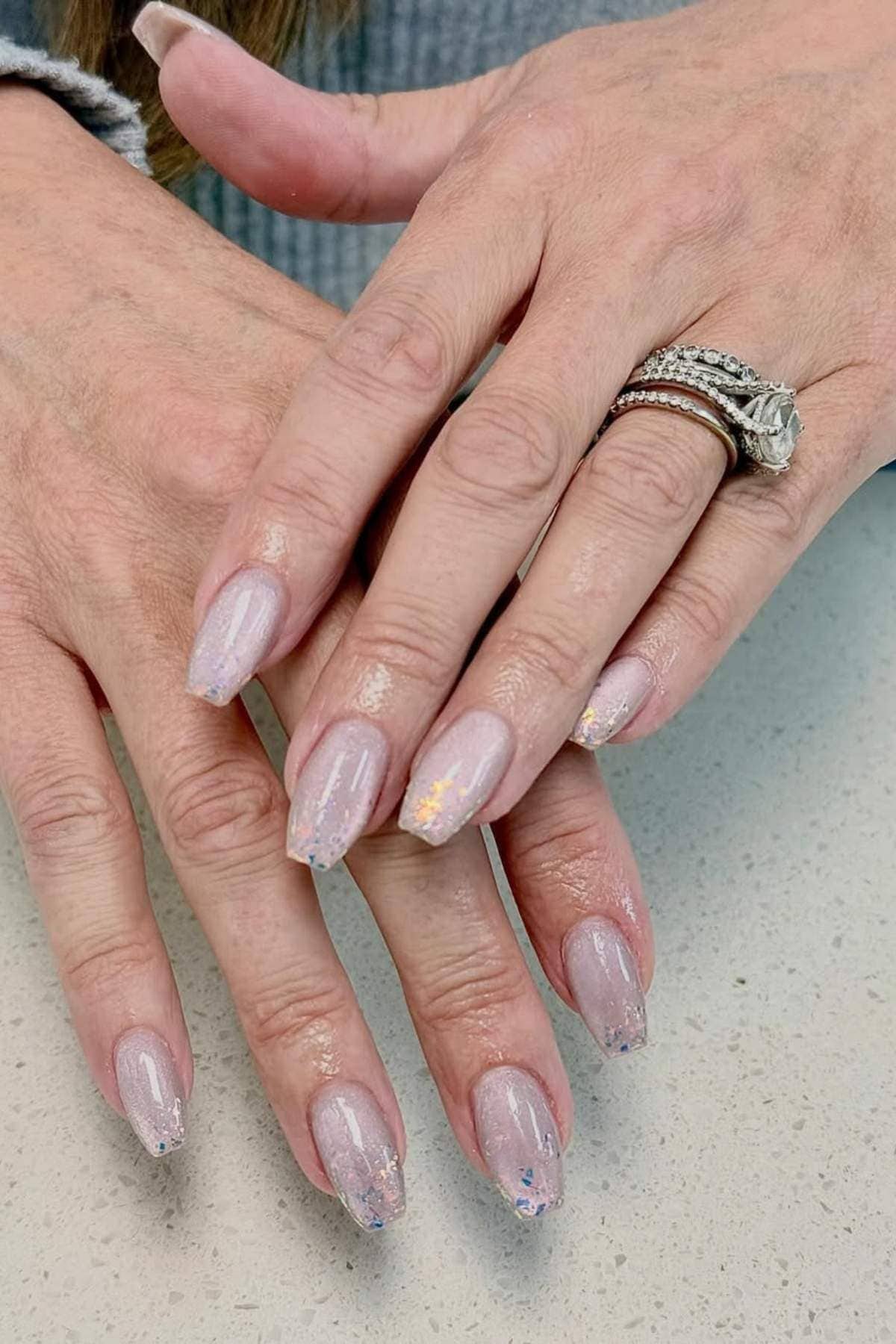 Close-up of hands with light pink, almond-shaped nails featuring subtle glitter tips; one hand has a diamond engagement and wedding ring.