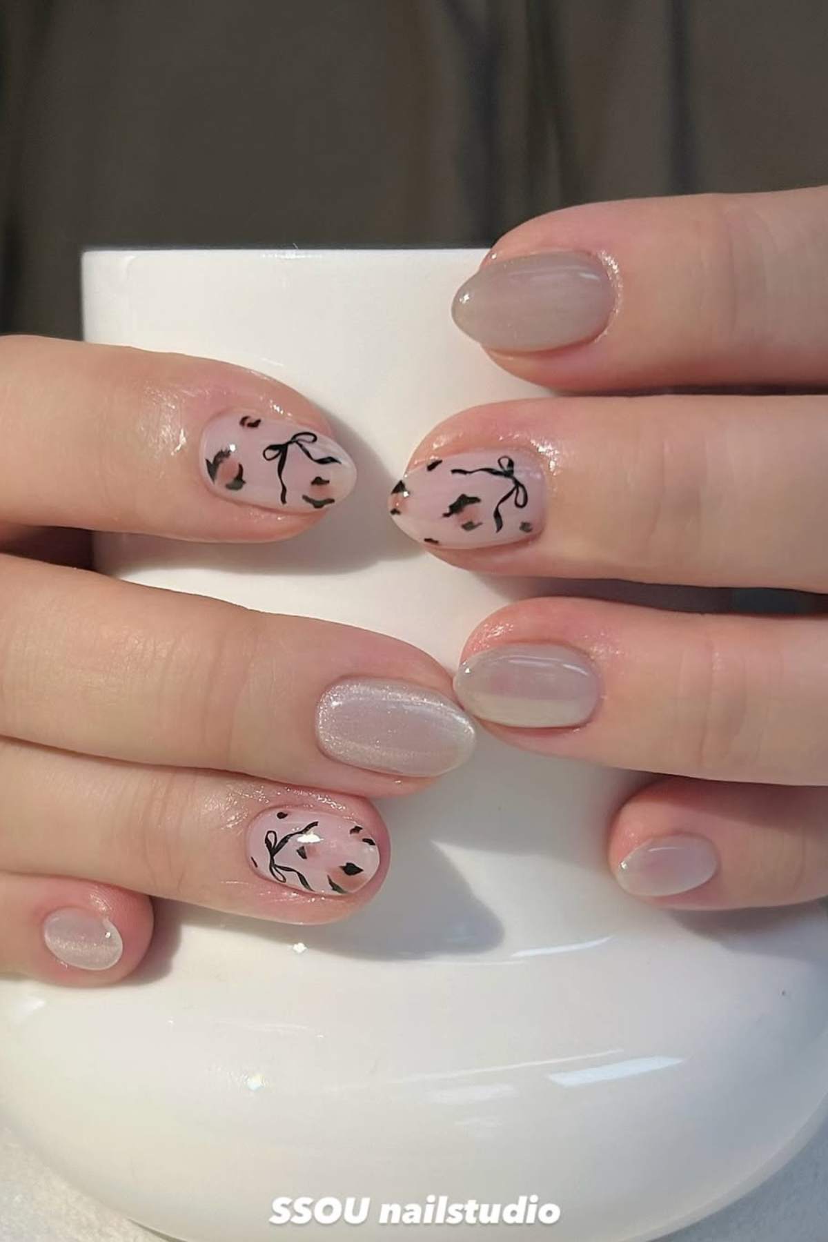 Hands with light pink manicured nails; some nails feature black abstract floral and leaf designs while others are painted with a sheer, shimmery polish.