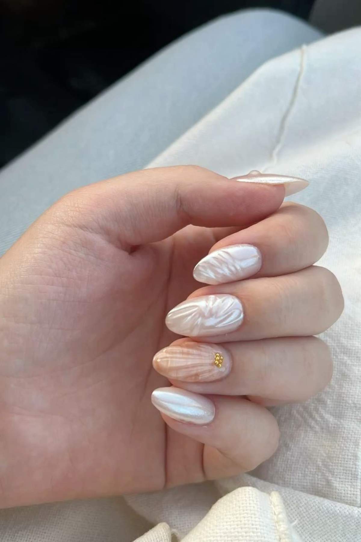 A hand with almond-shaped nails featuring a pearly white and pale peach marbled design, with a small gold bead on the ring finger nail.