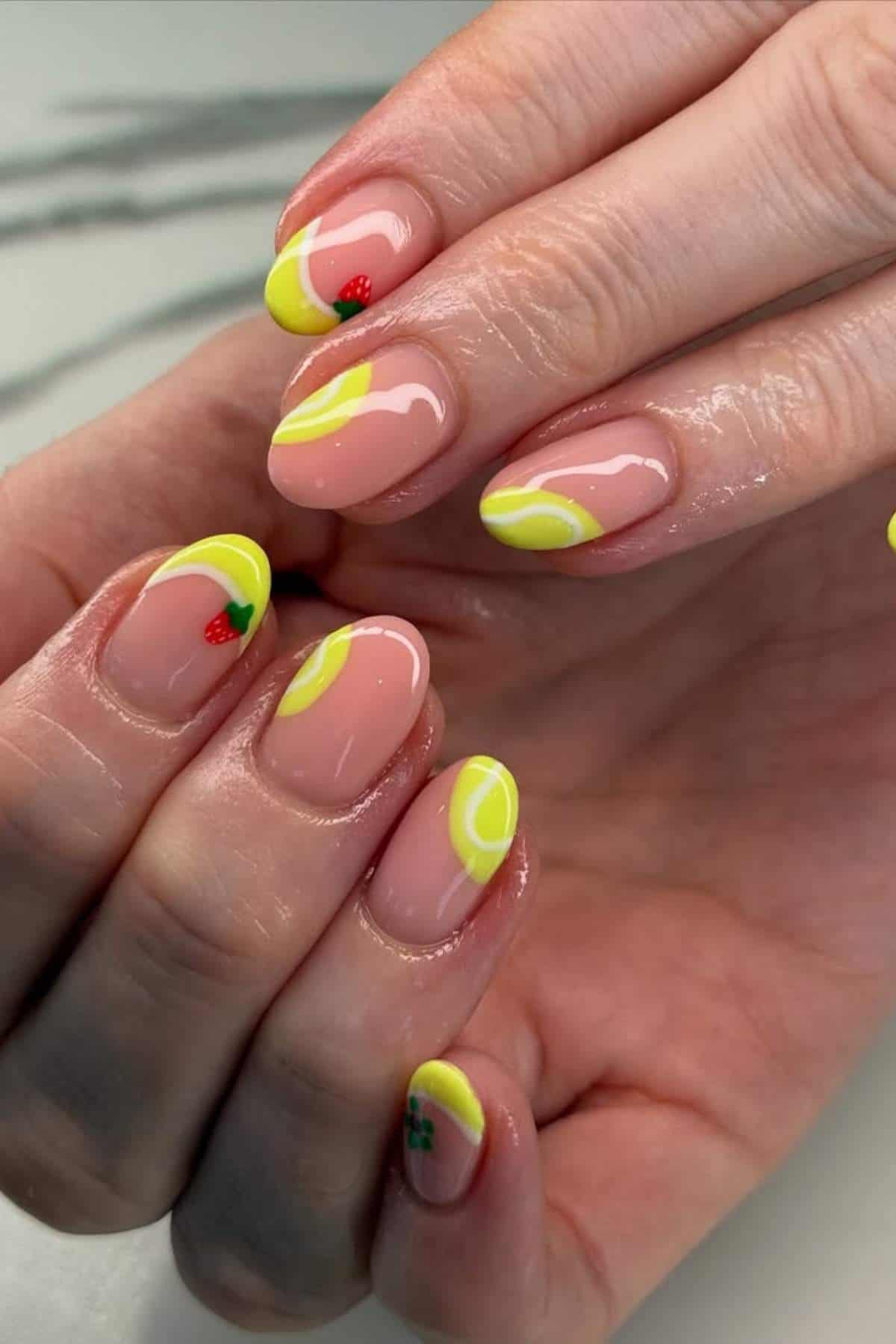 Hands with manicured nails featuring yellow French tips and small fruit designs, including strawberries and lemons, on a light background.