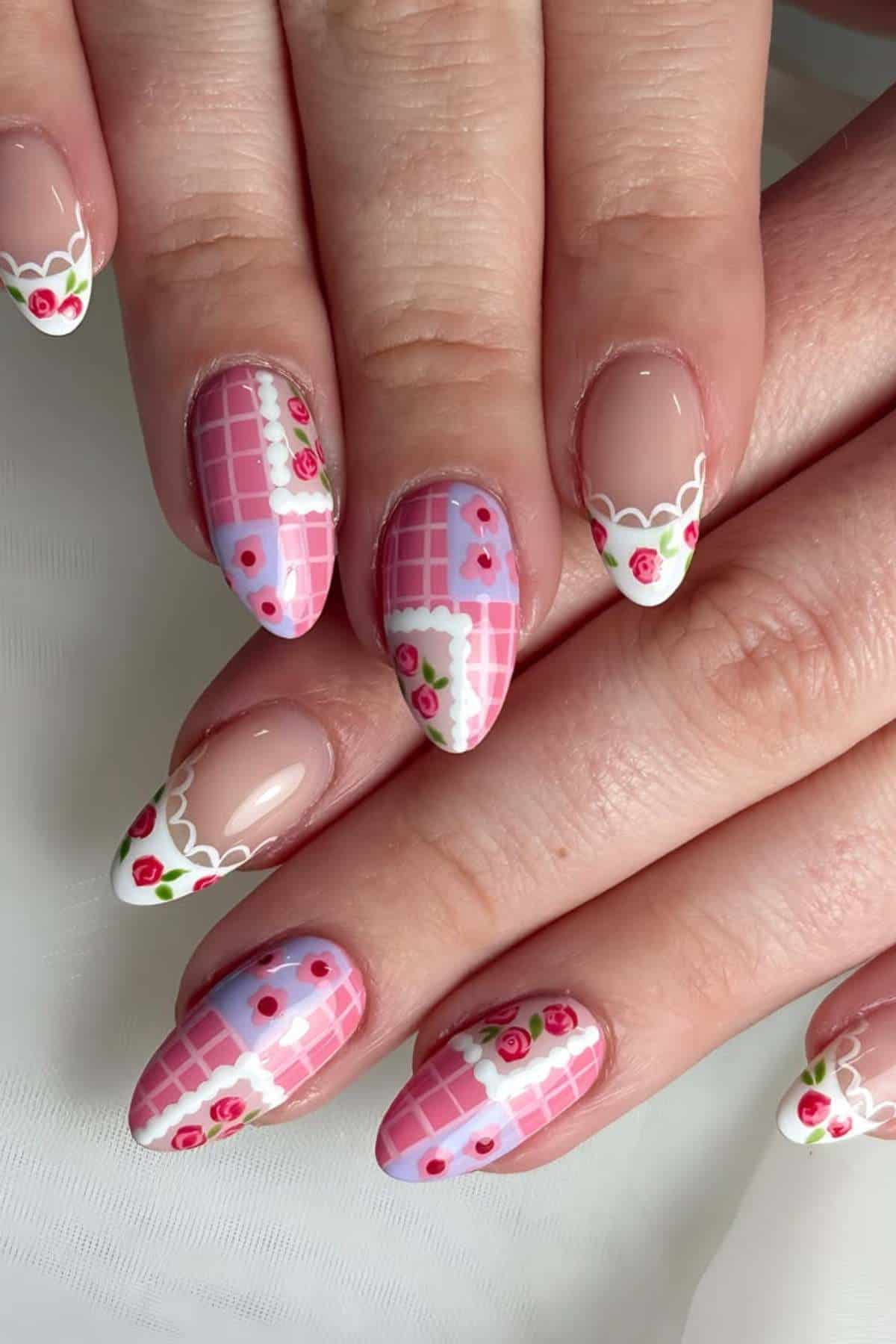 Hands with almond-shaped nails featuring pink and white checkered nail art, small red flowers, white scalloped edges, and rose designs on a glossy finish.