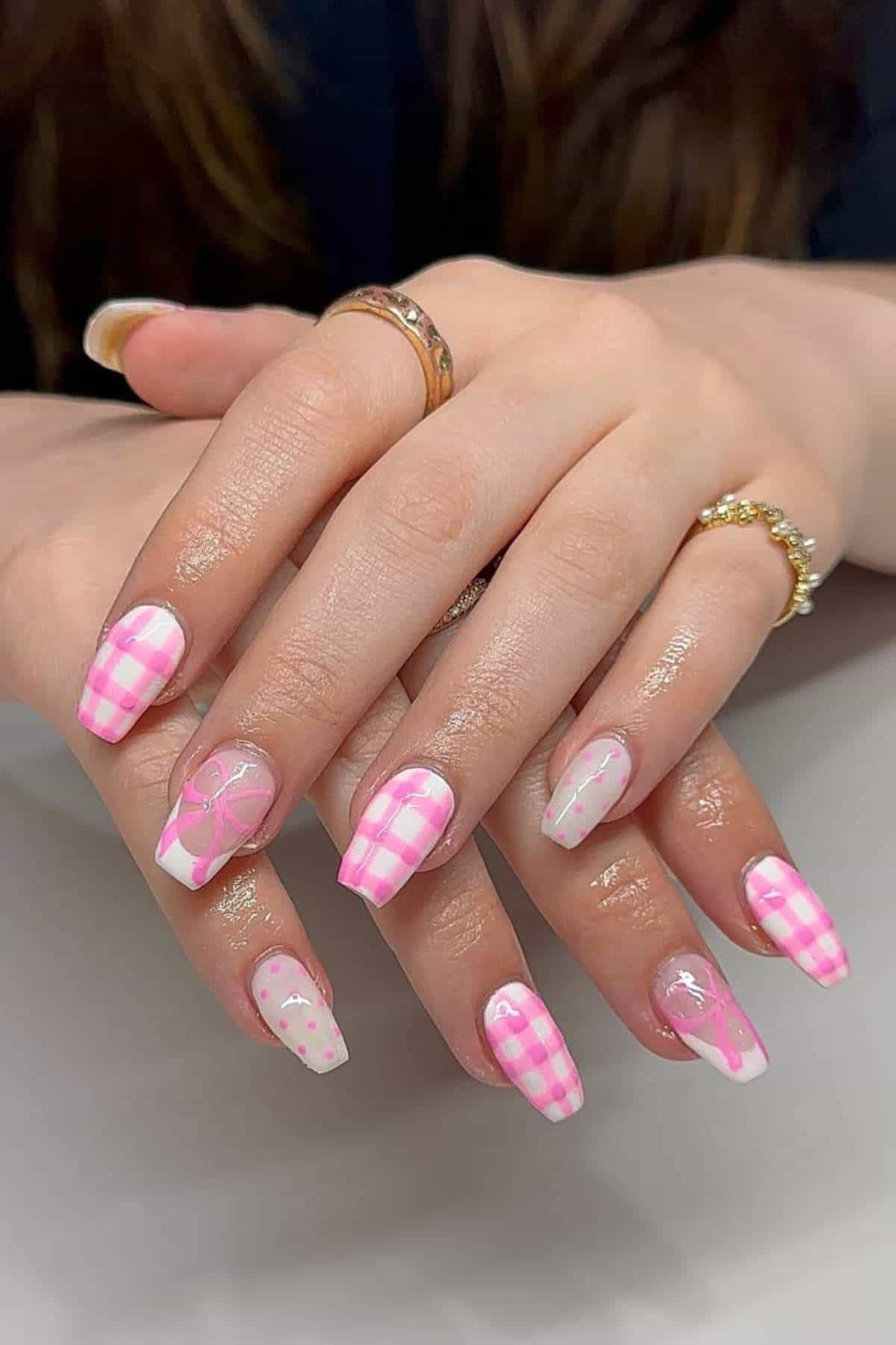 Hands with long, almond-shaped nails featuring pink and white checkered patterns, bows, dots, and hearts, displayed on a light surface.