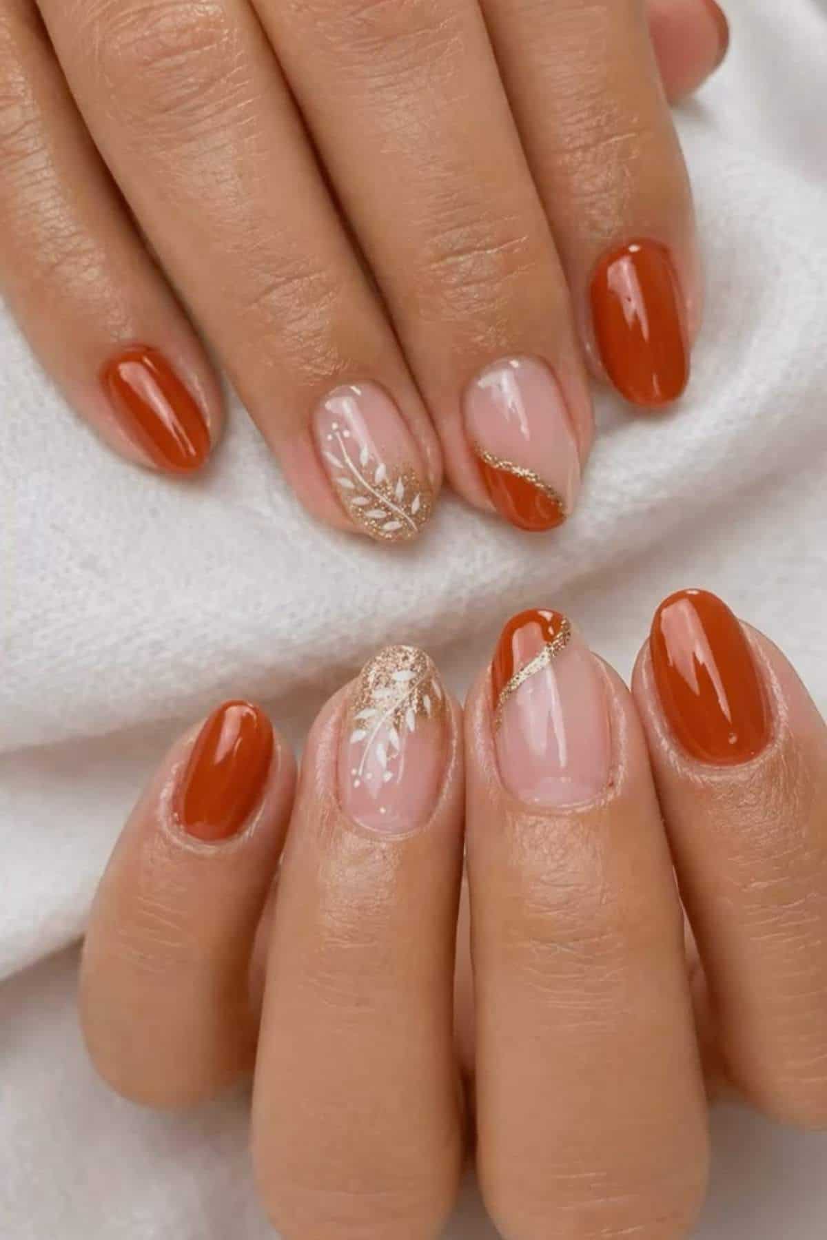 Hands with short, oval-shaped nails featuring burnt orange polish and accent nails with gold glitter, leaf designs, and diagonal gold lines on a neutral base.