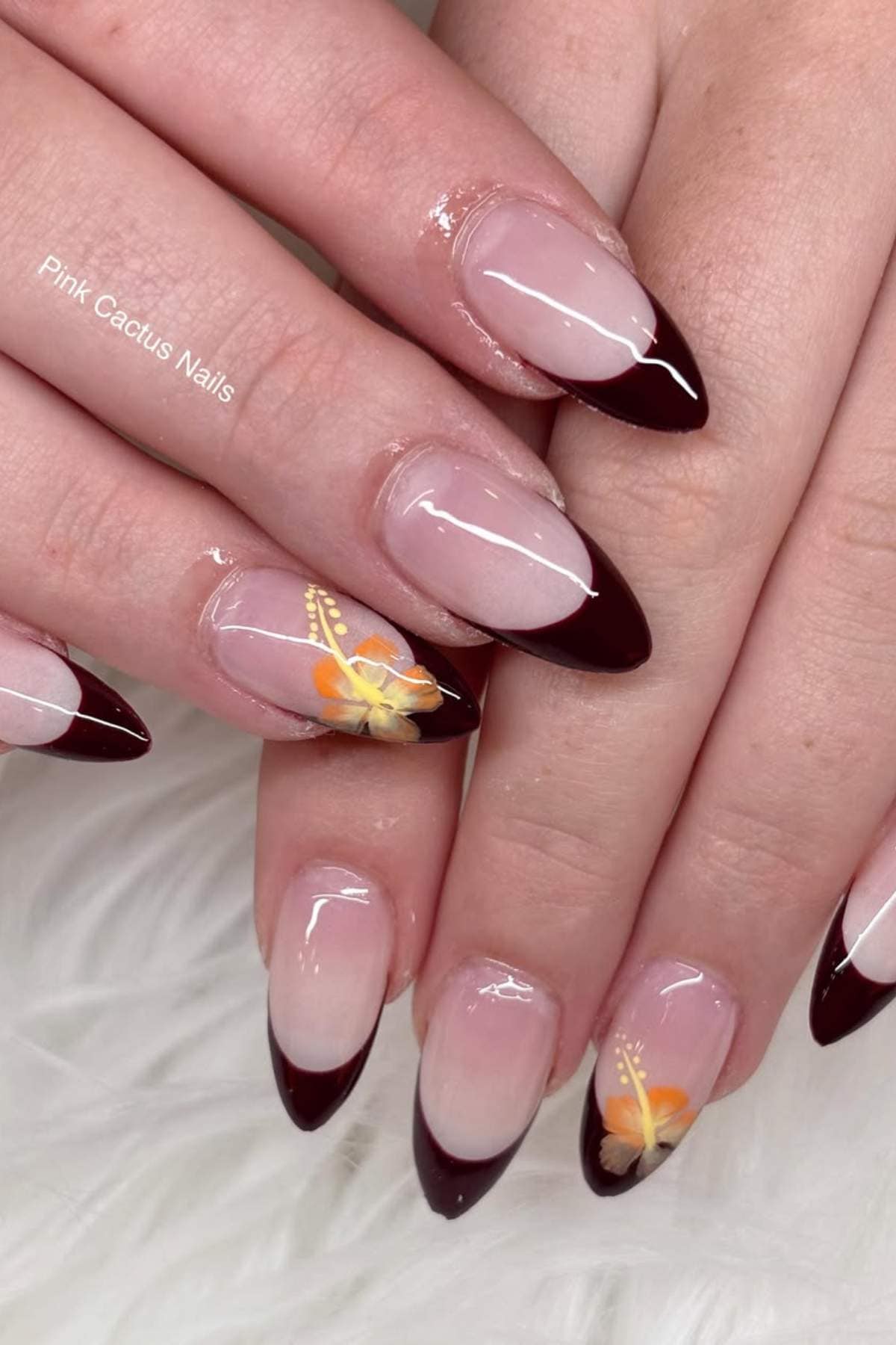 Hands with almond-shaped nails featuring dark burgundy French tips and two accent nails with yellow-orange butterfly nail art on a light, textured background.