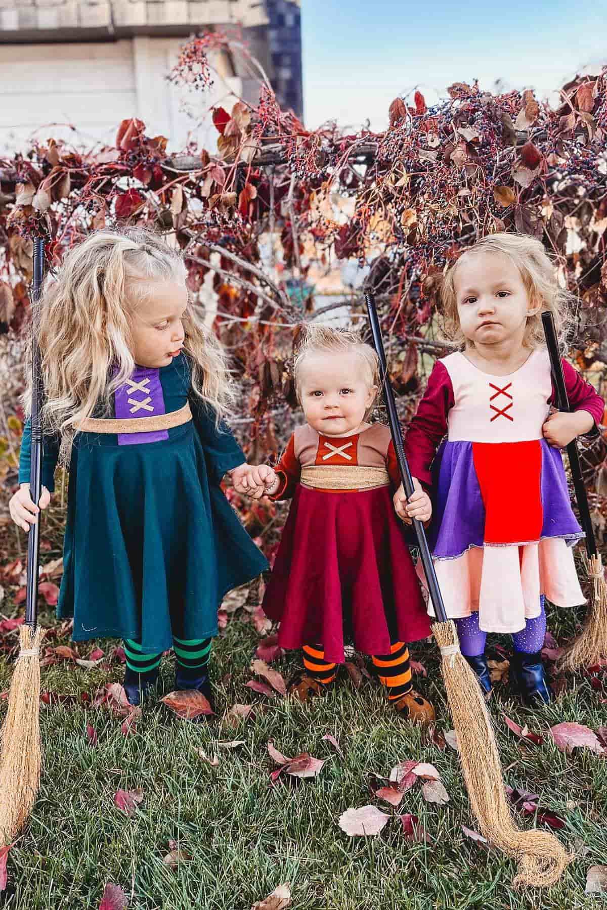 Three young children dressed in colorful witch costumes stand on grass holding brooms, with autumn leaves and vines in the background.