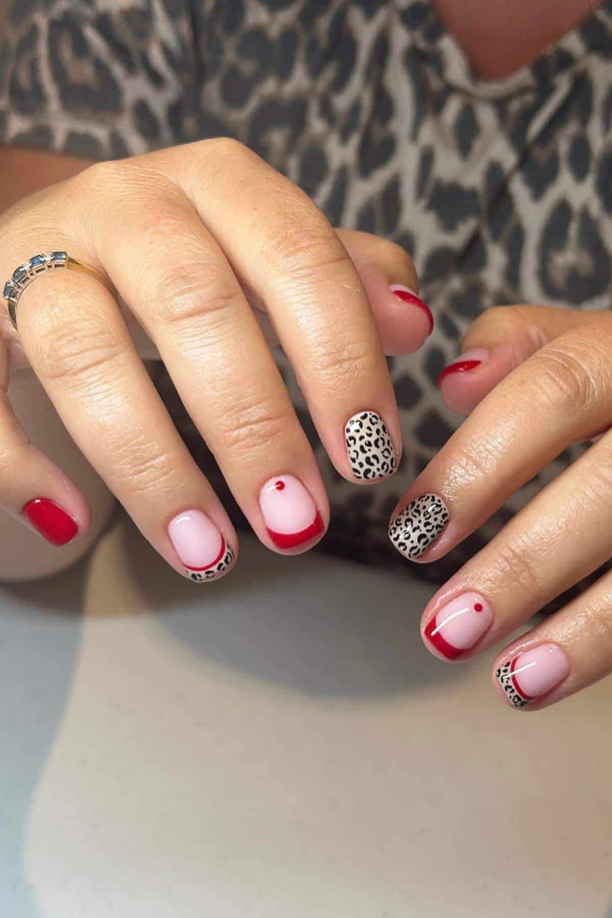 Hands with manicured nails featuring a mix of red polish, pink base with red tips and dots, and black-and-white leopard print designs, resting on a white surface.