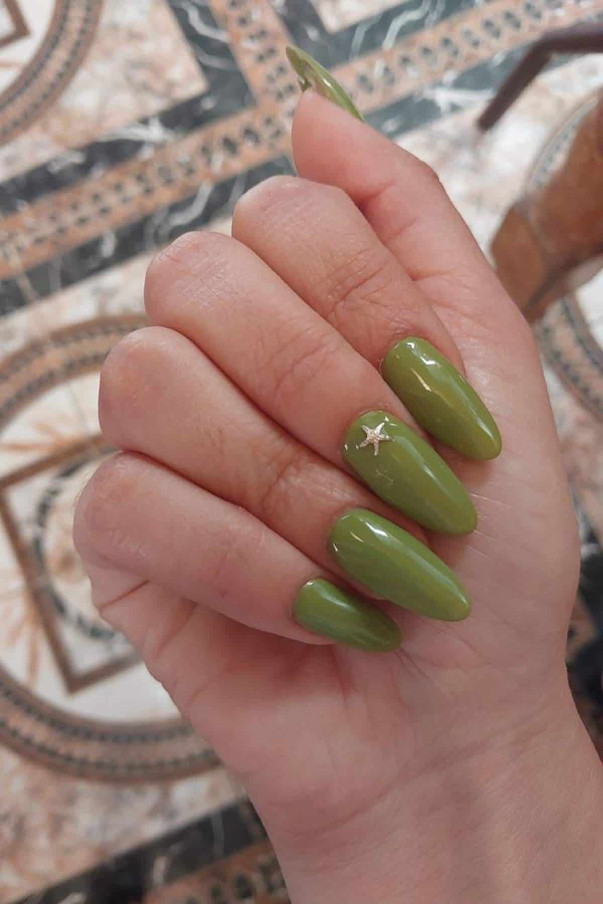 A hand with long, almond-shaped nails painted green; one nail features a small silver star decoration.