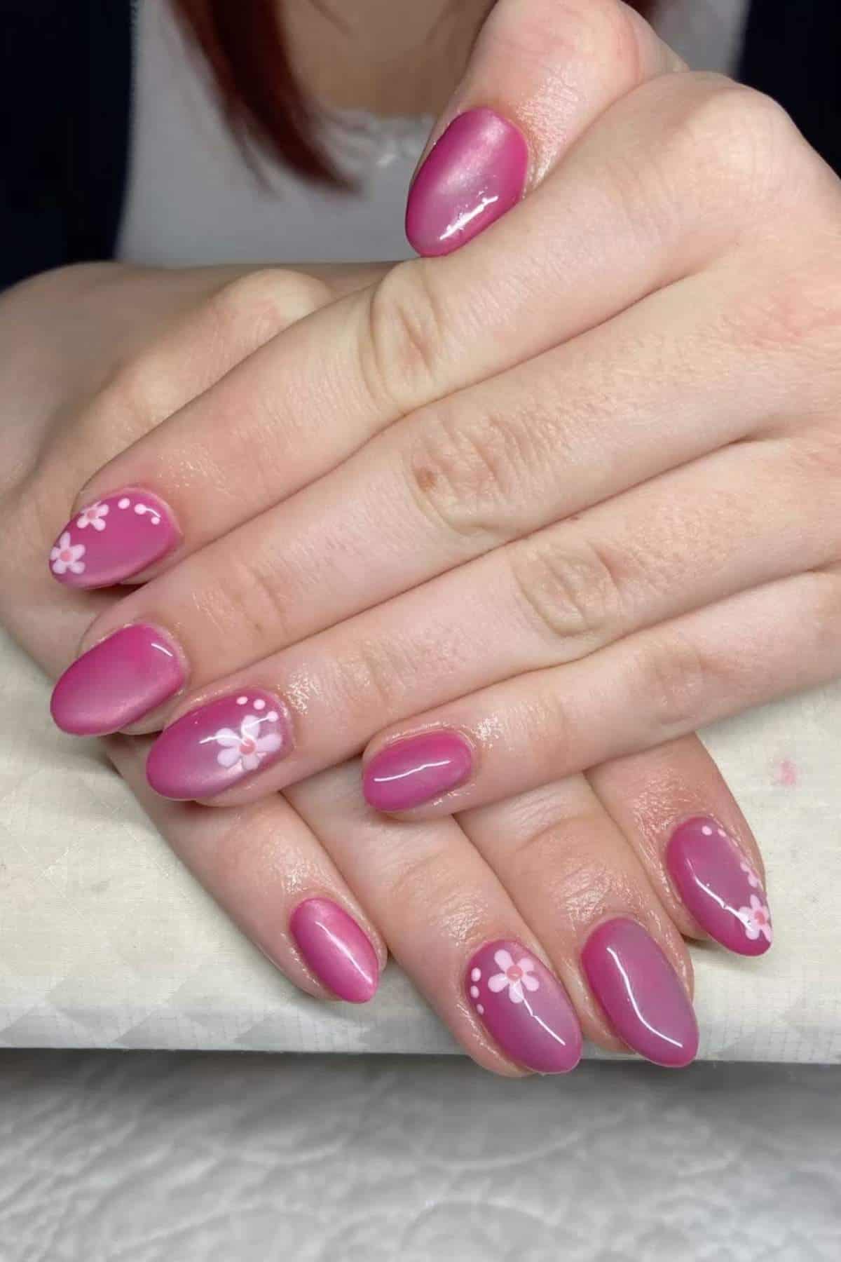 Hands with short, almond-shaped nails painted pink, featuring white flower designs and dotted accents on two nails.