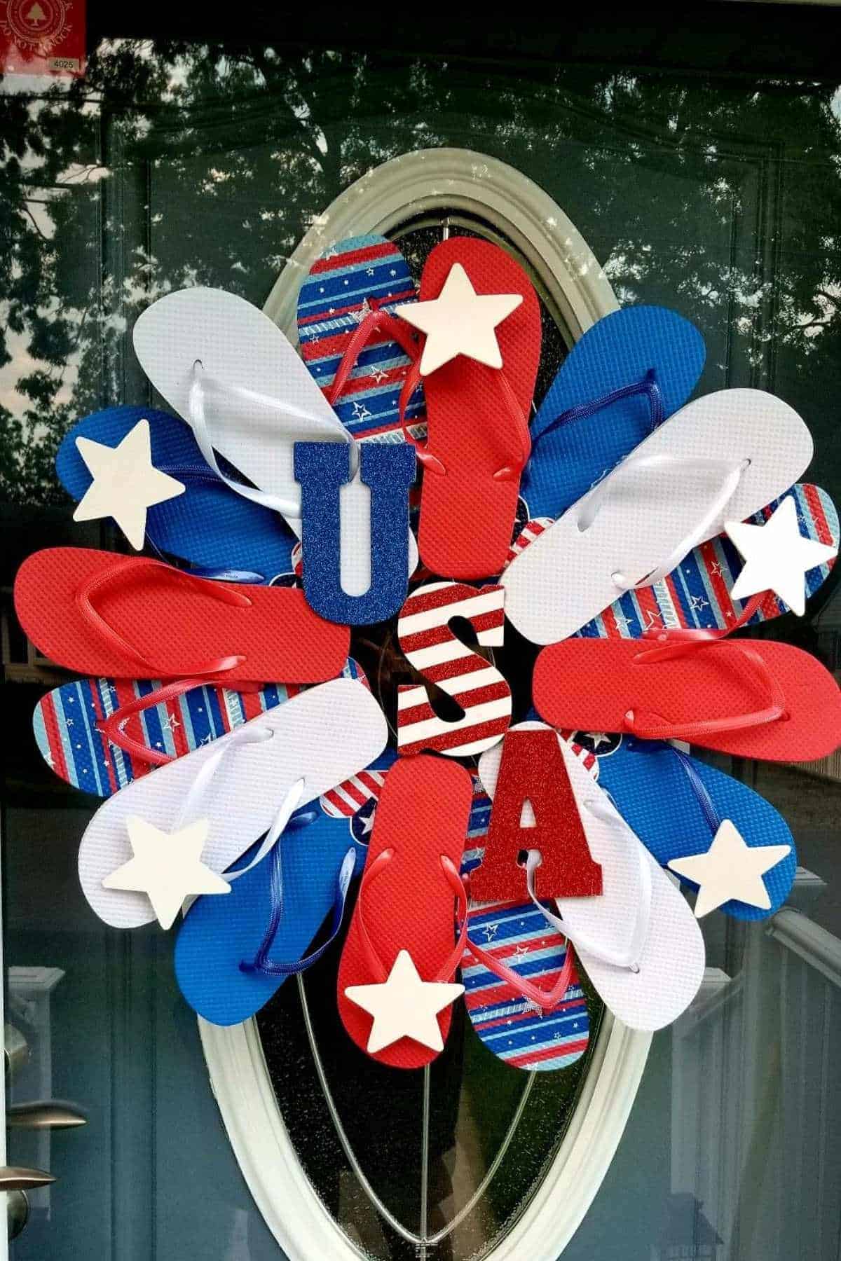 A wreath made of red, white, and blue flip-flops, decorated with stars and the letters "USA," is displayed on a glass door.