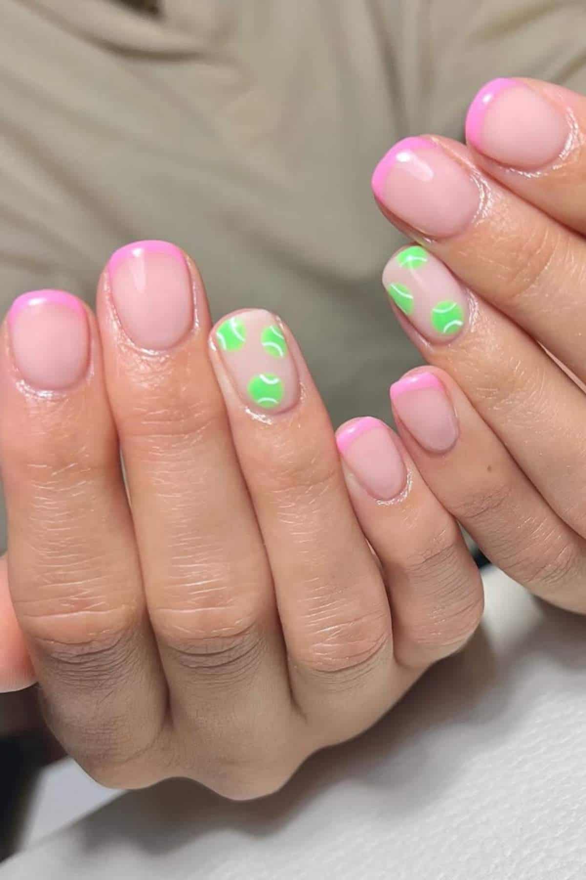 Hands with short, natural-shaped nails featuring pink French tips and two accent nails with green abstract dot designs on a neutral base.