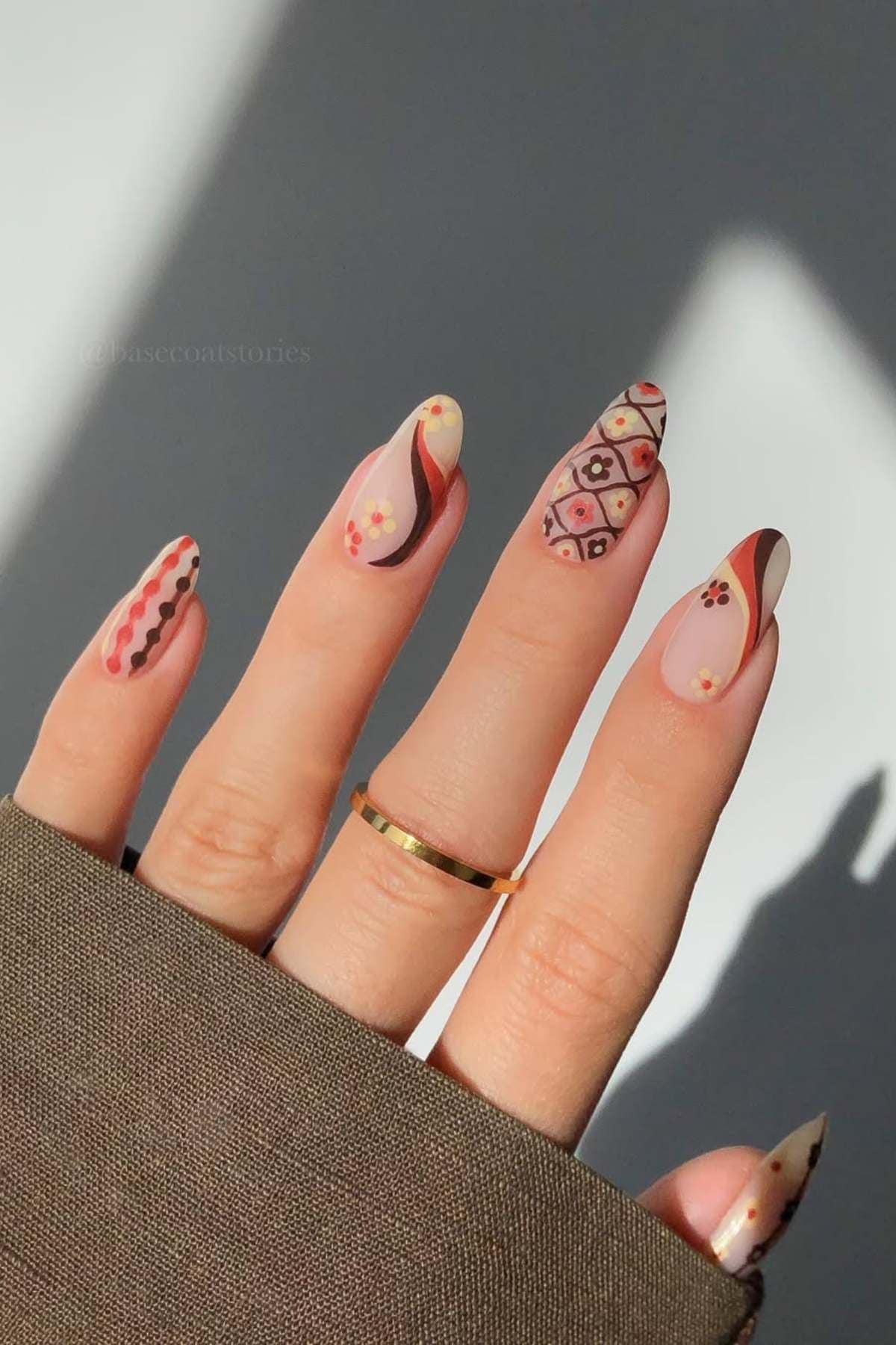 A hand with long almond-shaped nails features intricate nail art with geometric, floral, and wavy patterns in brown, beige, and red tones. The person wears a gold ring and a brown sleeve.
