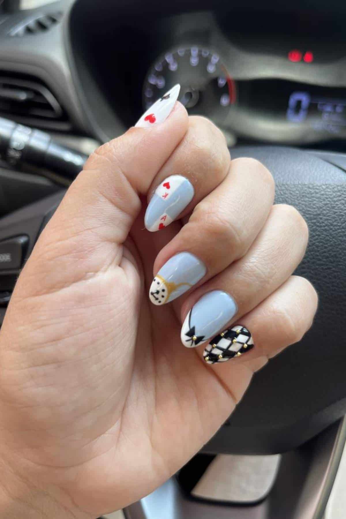 A hand with light blue nail polish featuring casino-themed nail art, including playing cards, a poker chip, dice, and a diamond pattern, is shown in front of a car steering wheel.
