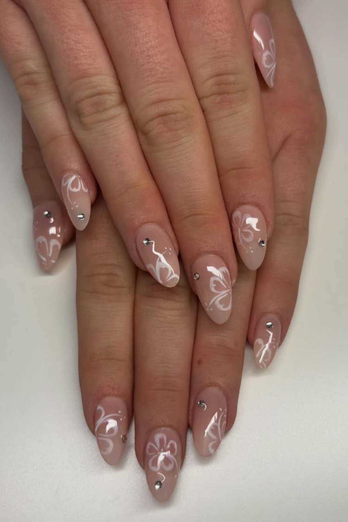 Hands with almond-shaped nails featuring a pale pink base, white floral designs, and small rhinestone accents on each nail.