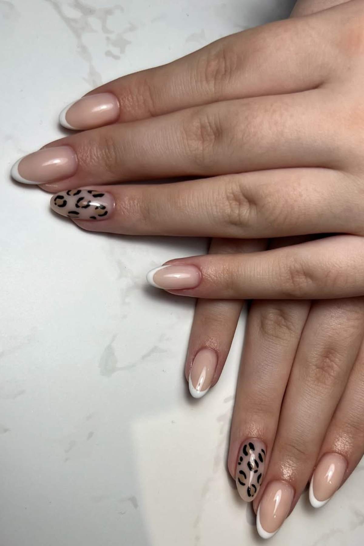 Hands with manicured nails featuring a nude base, white French tips, and two accent nails with black and brown leopard print design.
