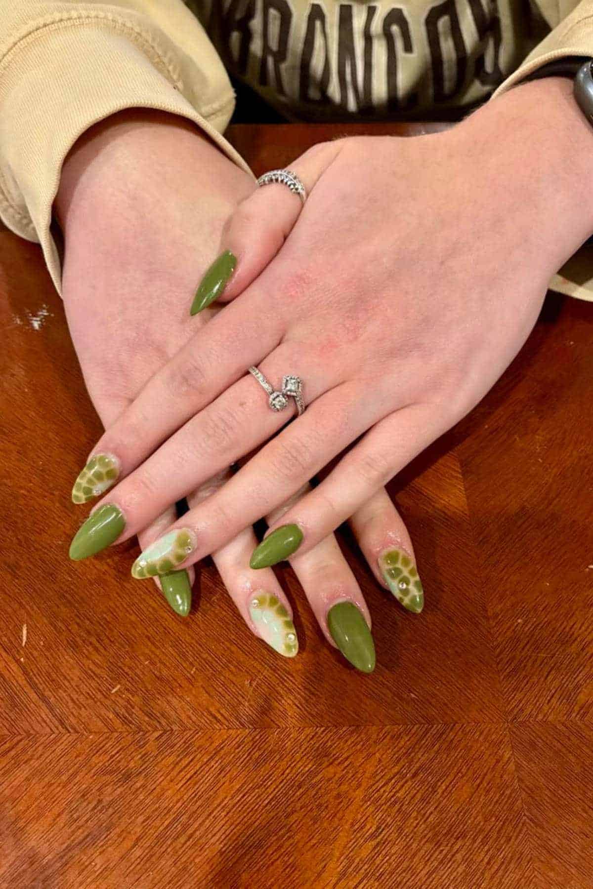 Hands with long, almond-shaped green nails featuring a mix of solid color and tortoiseshell-like accent designs, resting on a wooden surface.