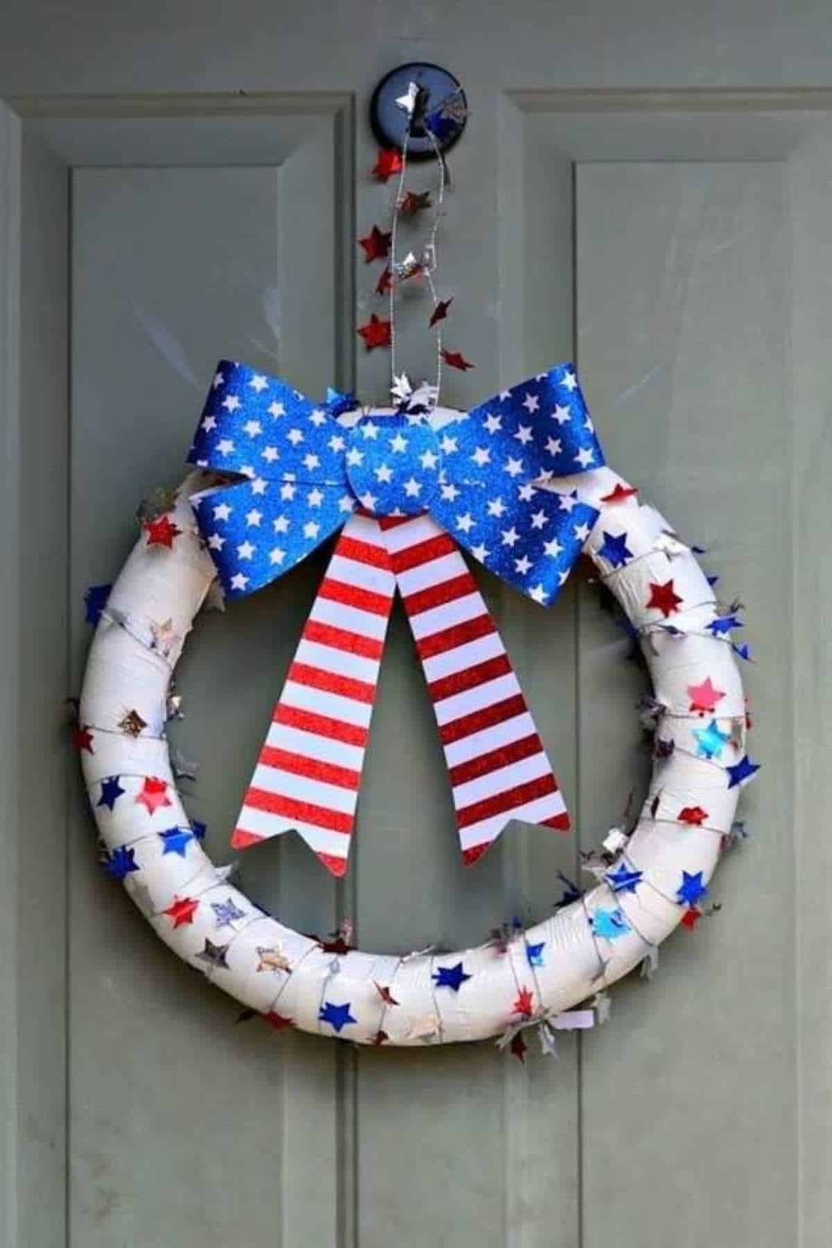 A wreath decorated with a large blue, white, and red bow featuring stars and stripes, and wrapped with star-shaped garland, hanging on a gray door.