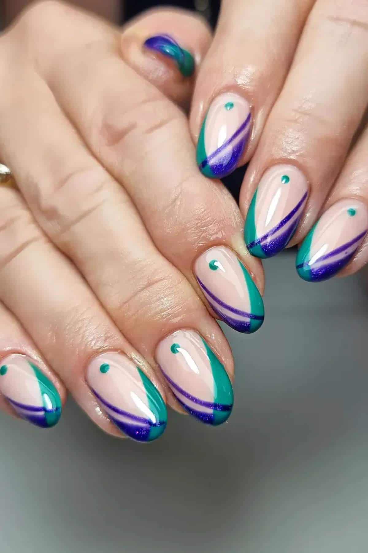 Close-up of hands with almond-shaped nails painted with teal and purple diagonal tips, clear base, and a small teal dot near the cuticle on each nail.