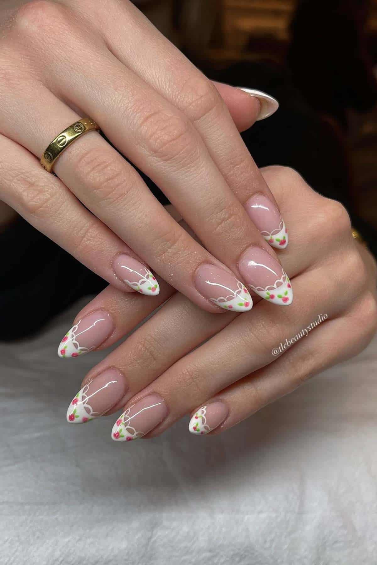 Hands with almond-shaped nails featuring a clear base and white French tips decorated with small yellow and green floral designs. One hand has a gold ring.