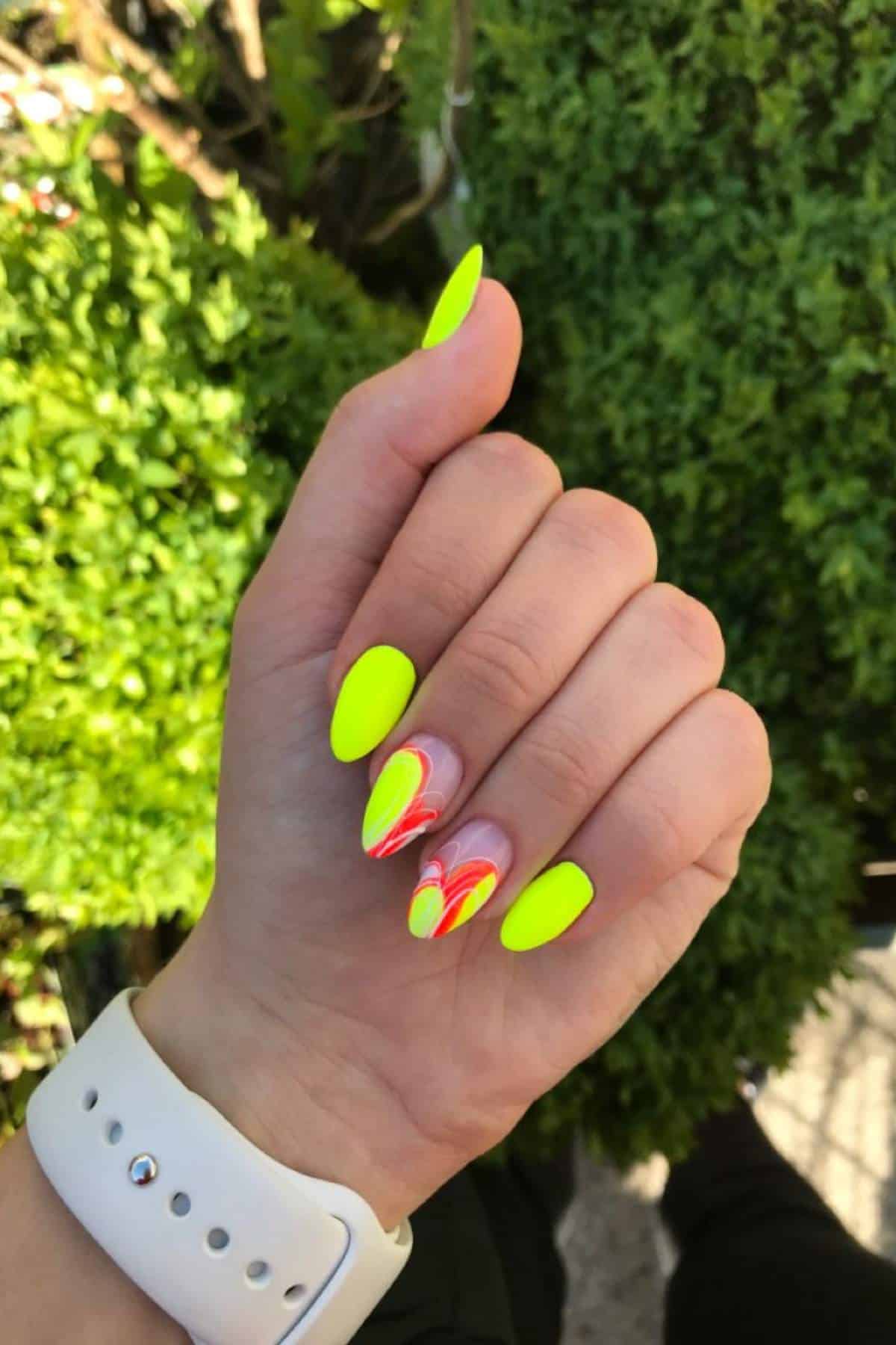 A hand with long, almond-shaped nails painted neon yellow, with two accent nails featuring abstract red, yellow, and white swirl designs, against a leafy green background.