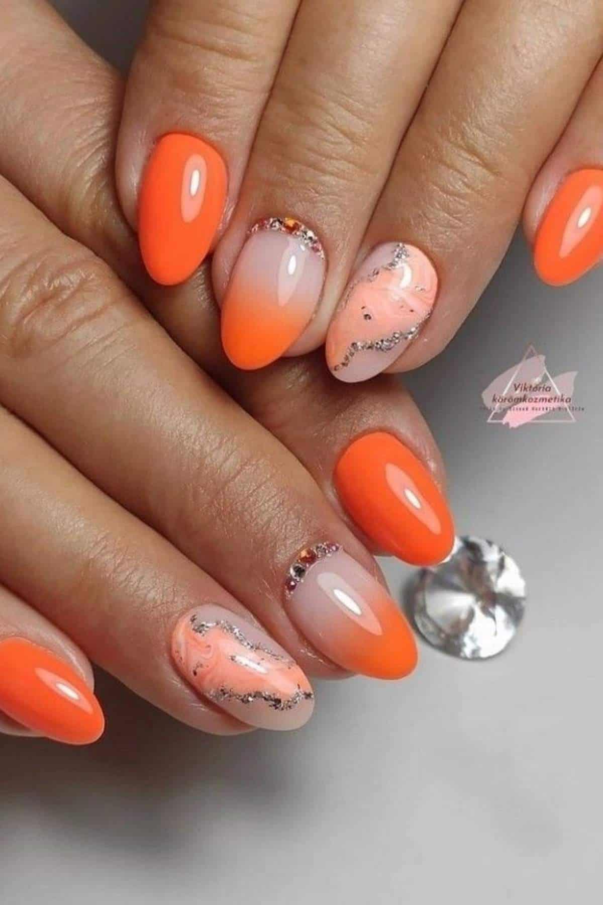 Hands with almond-shaped nails featuring a bright orange polish, marble accent nails with orange and silver glitter, and small rhinestone decorations.