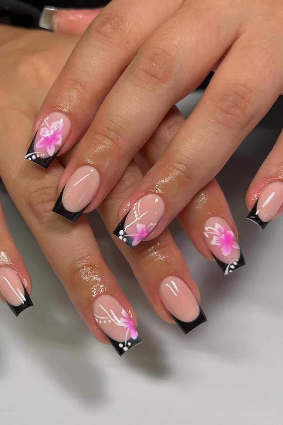 A hand with long, square-shaped nails featuring black French tips and pink floral designs with white accents on a glossy nude base.