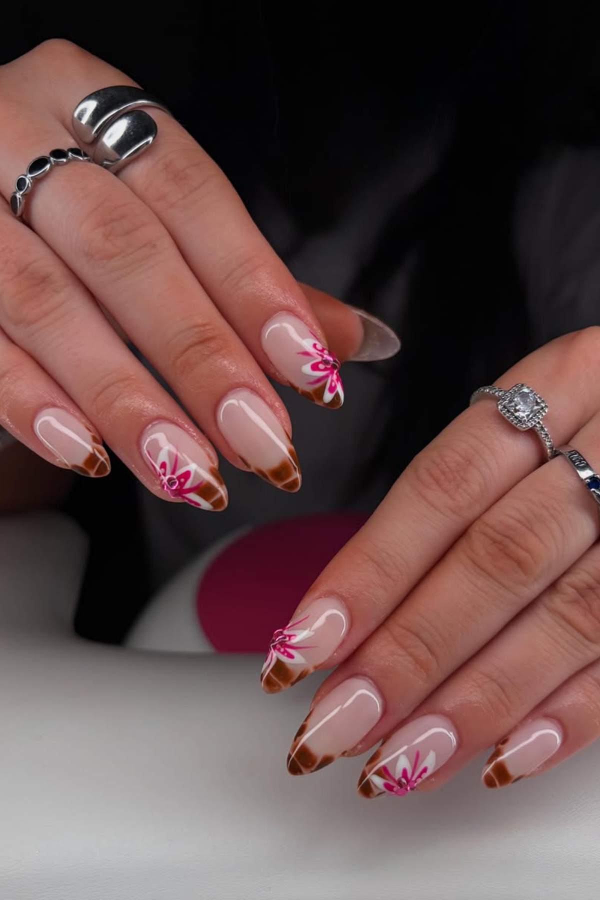 Hands with almond-shaped nails featuring brown French tips and pink floral designs. The person is wearing silver rings on several fingers.