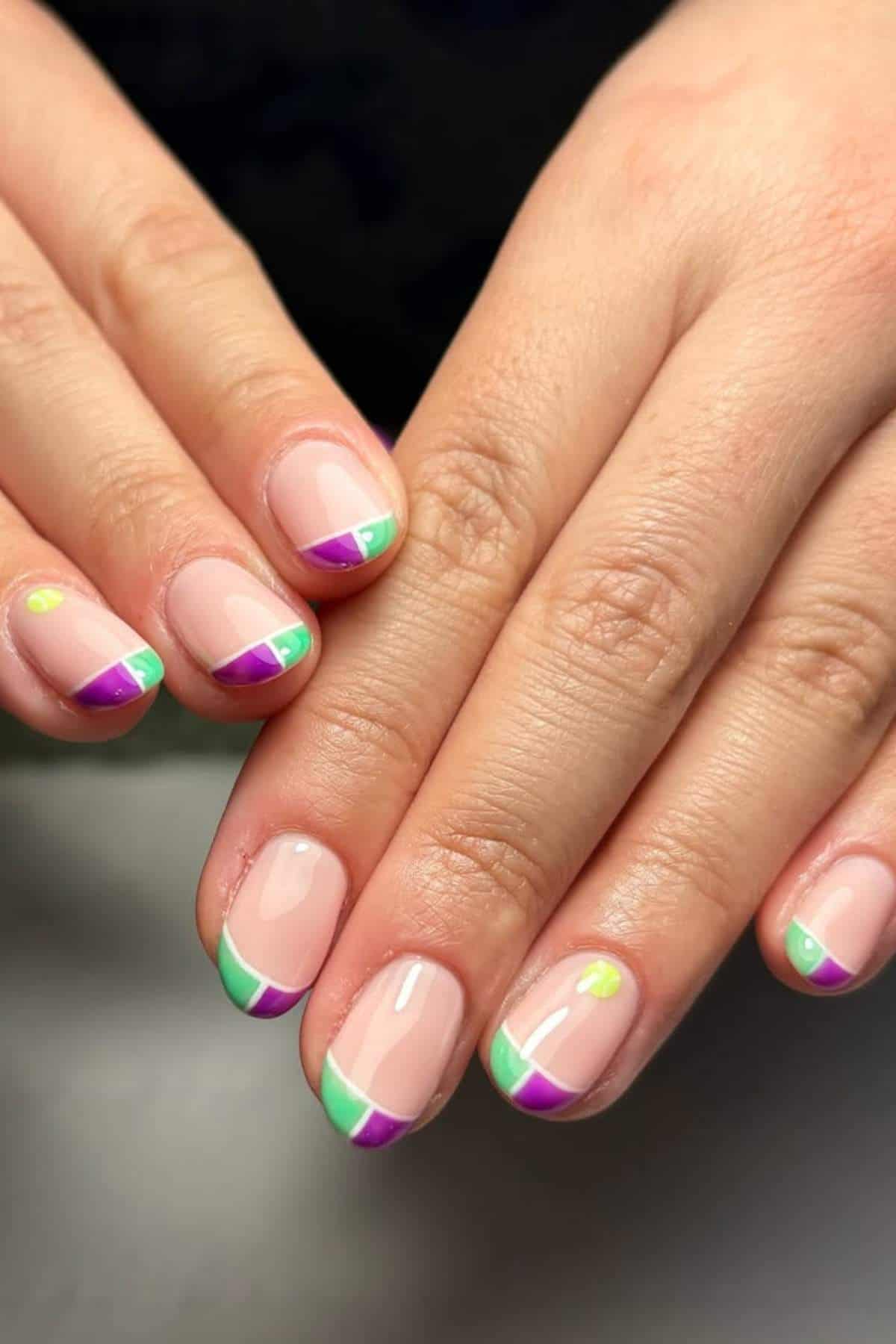 Close-up of hands with short nails featuring a colorful French tip manicure with green, purple, yellow, and pink accents on a nude base.