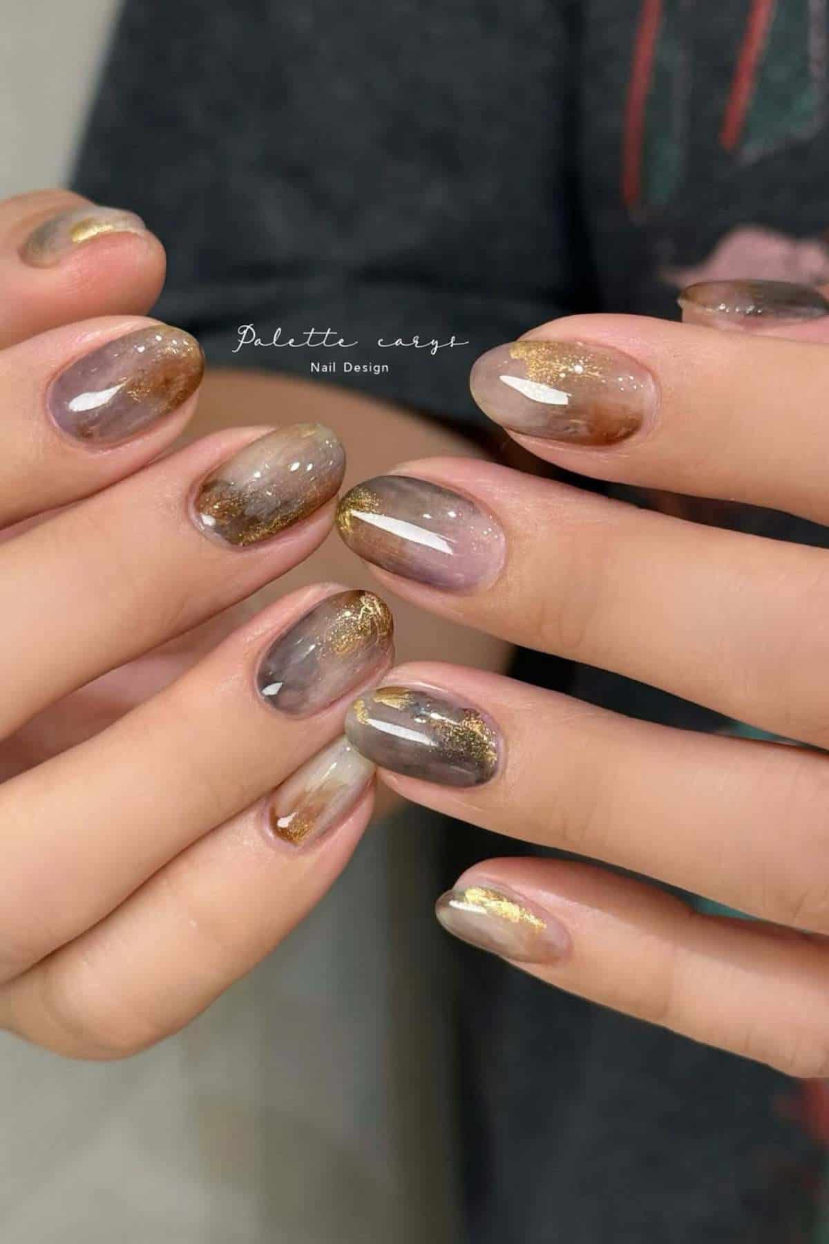 Hands with short, oval-shaped nails featuring a translucent brown and gray marble design with gold accents.