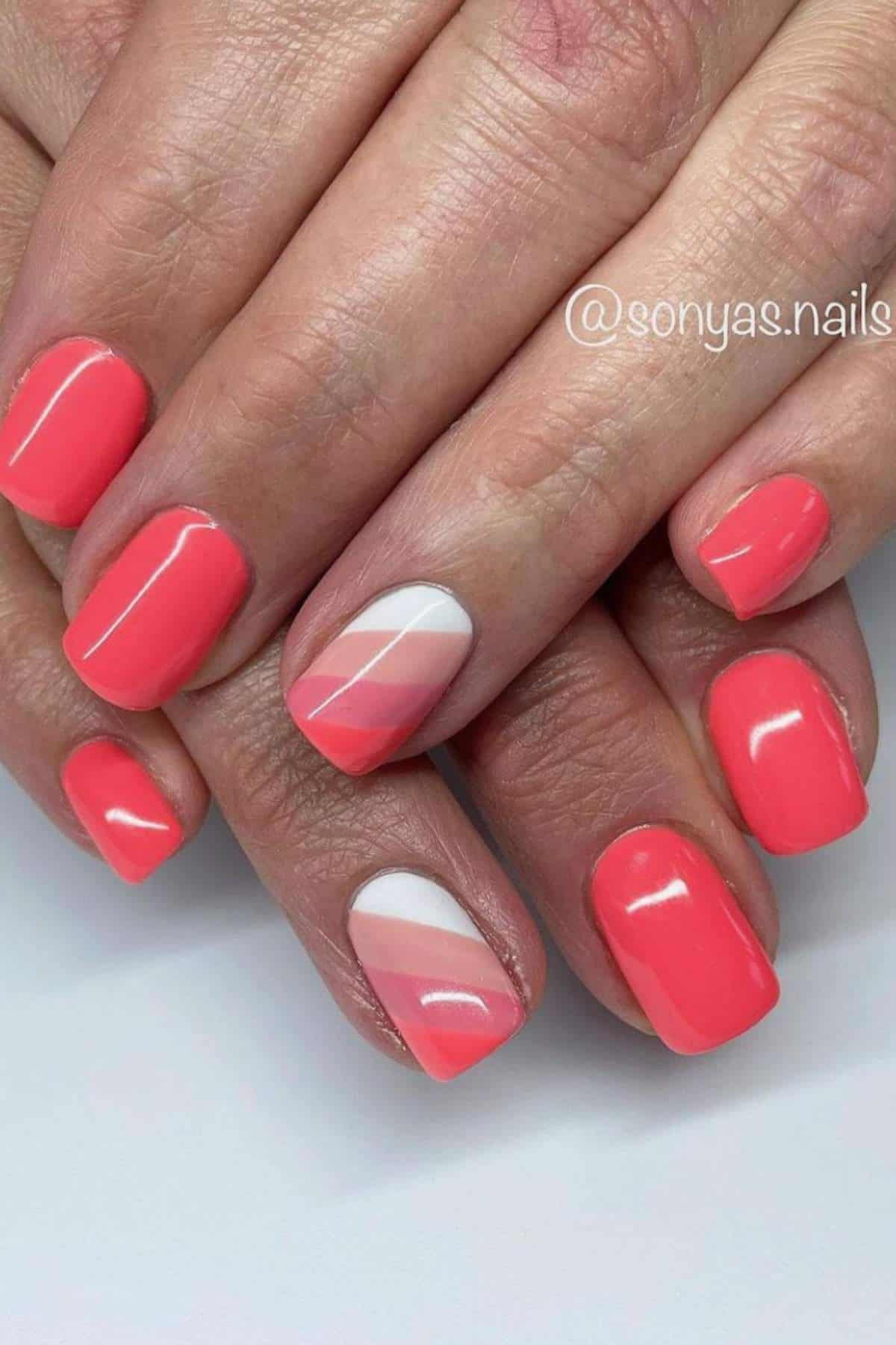 Close-up of hands with short, square-shaped nails painted in glossy coral pink, and accent nails featuring geometric white and pink diagonal lines.