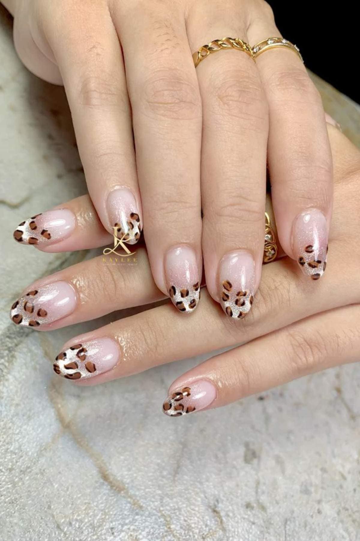 Hands with manicured nails featuring a glossy nude base and brown leopard print design on the tips.