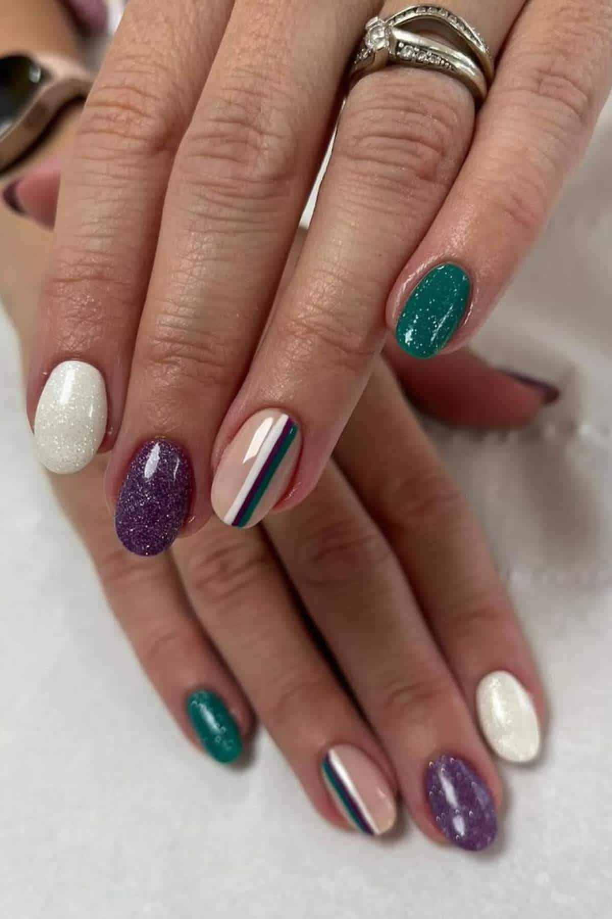 Hands with manicured nails painted in various colors including white, purple glitter, teal, and a nude base with multicolored stripes; person wears a ring on one finger.