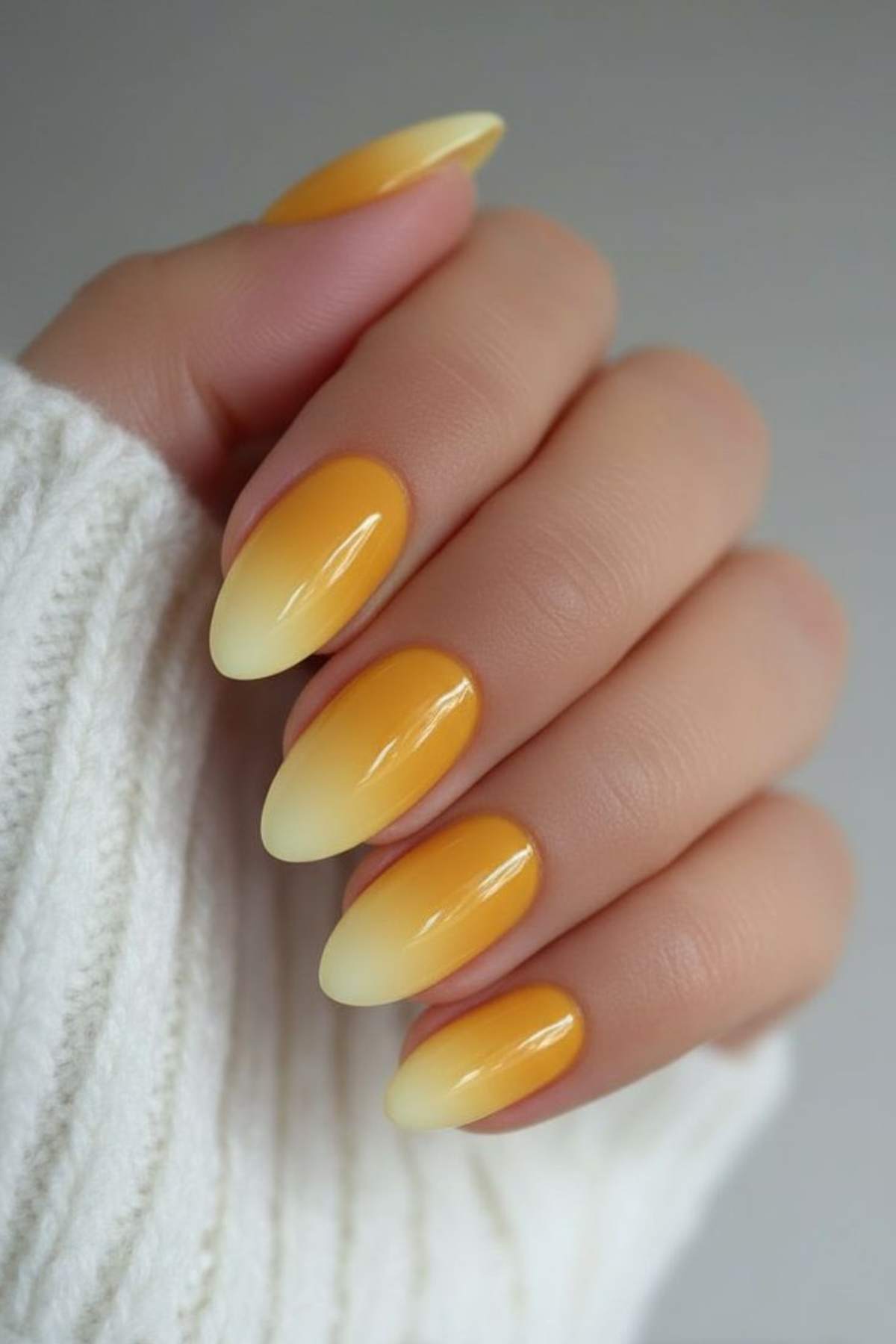 A hand with almond-shaped nails painted in a yellow and orange ombre design, shown against a white knitted sweater background.