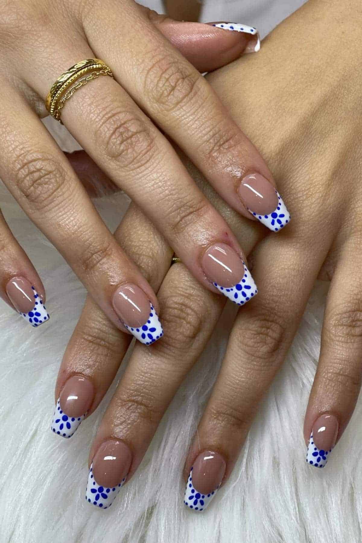 Hands with ballerina-shaped nails featuring a French tip design; the tips are white with blue polka dots. The person is wearing gold rings. Nails rest on a white textured surface.