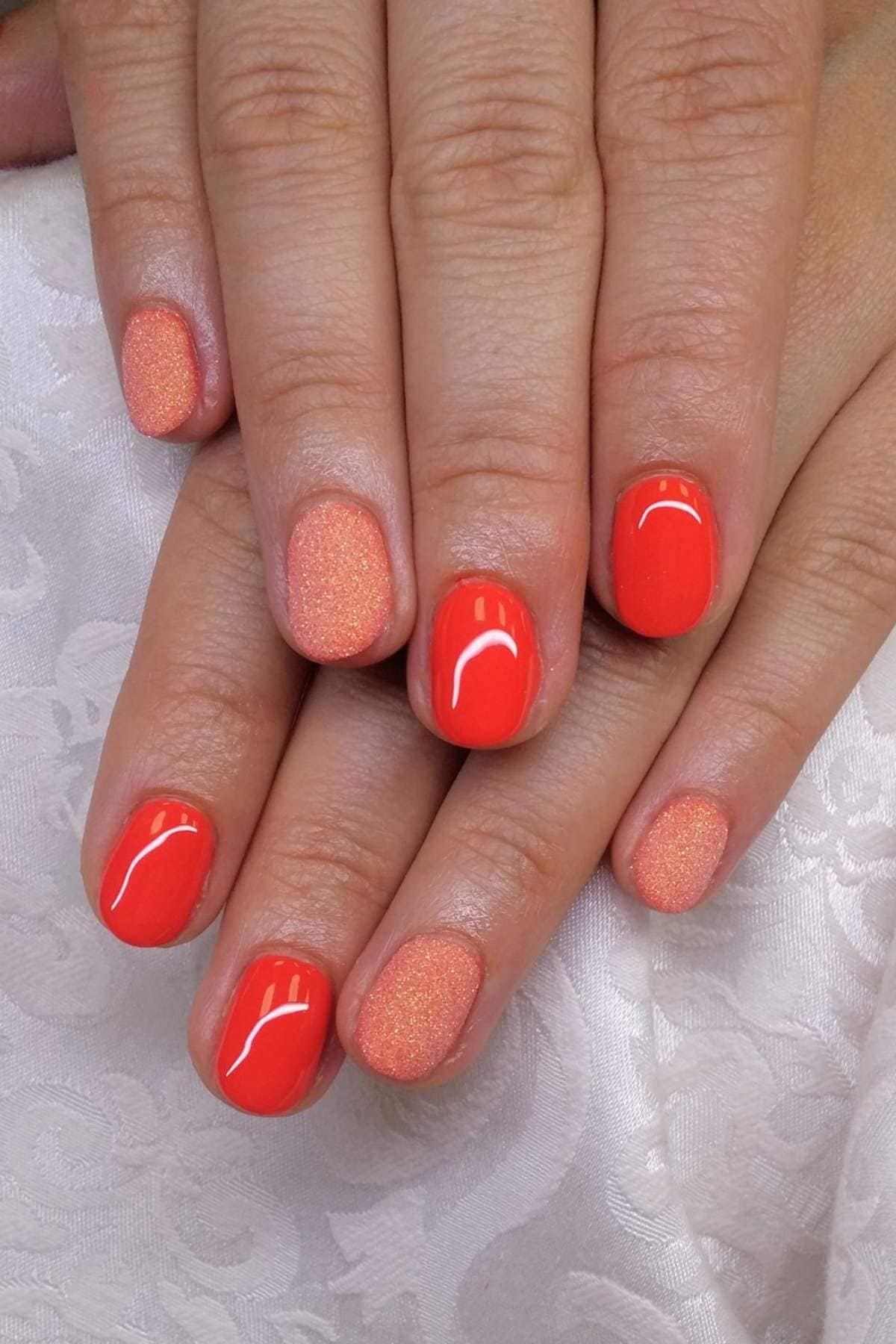 Close-up of hands with short, rounded nails painted in glossy coral orange polish, with two accent nails featuring a subtle shimmer effect.