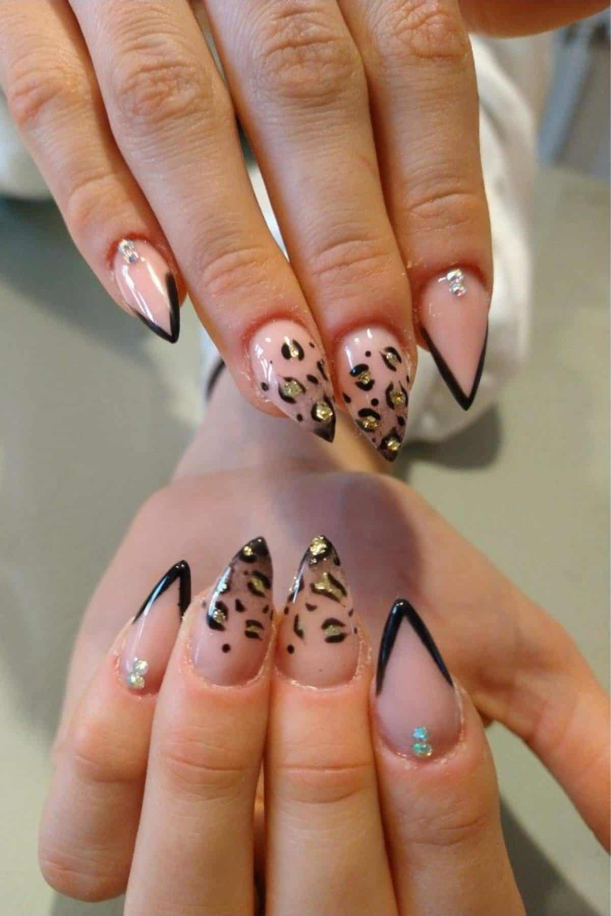 Hands with long, pointed nails featuring black tips, leopard print designs, and small rhinestone accents on a nude base.