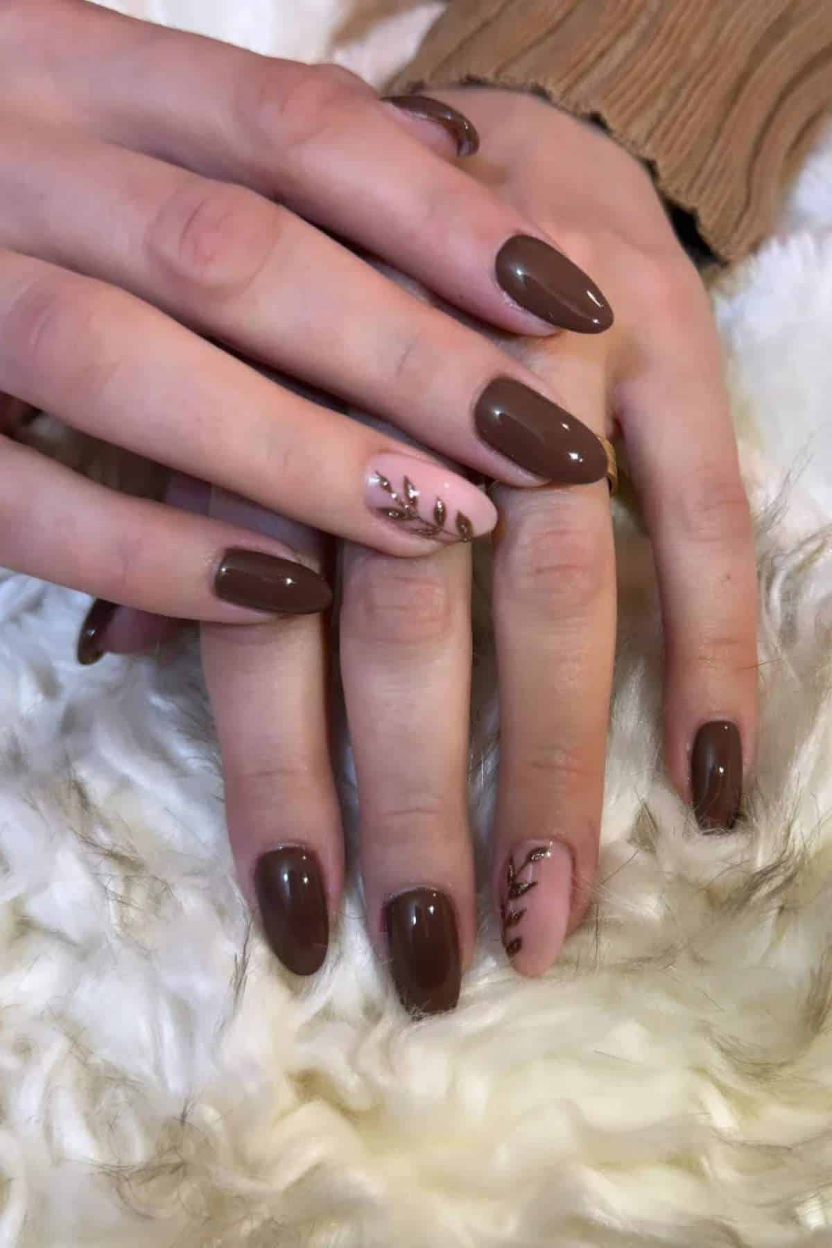 Hands with almond-shaped nails painted brown, with accent nails in light pink featuring a leaf design, resting on a fuzzy white surface.