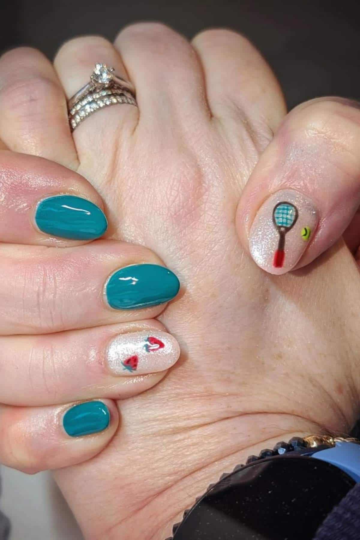 A hand with teal and glittery nail polish, featuring small painted designs of tennis rackets and balls on two nails. The person is wearing a ring and a smartwatch.