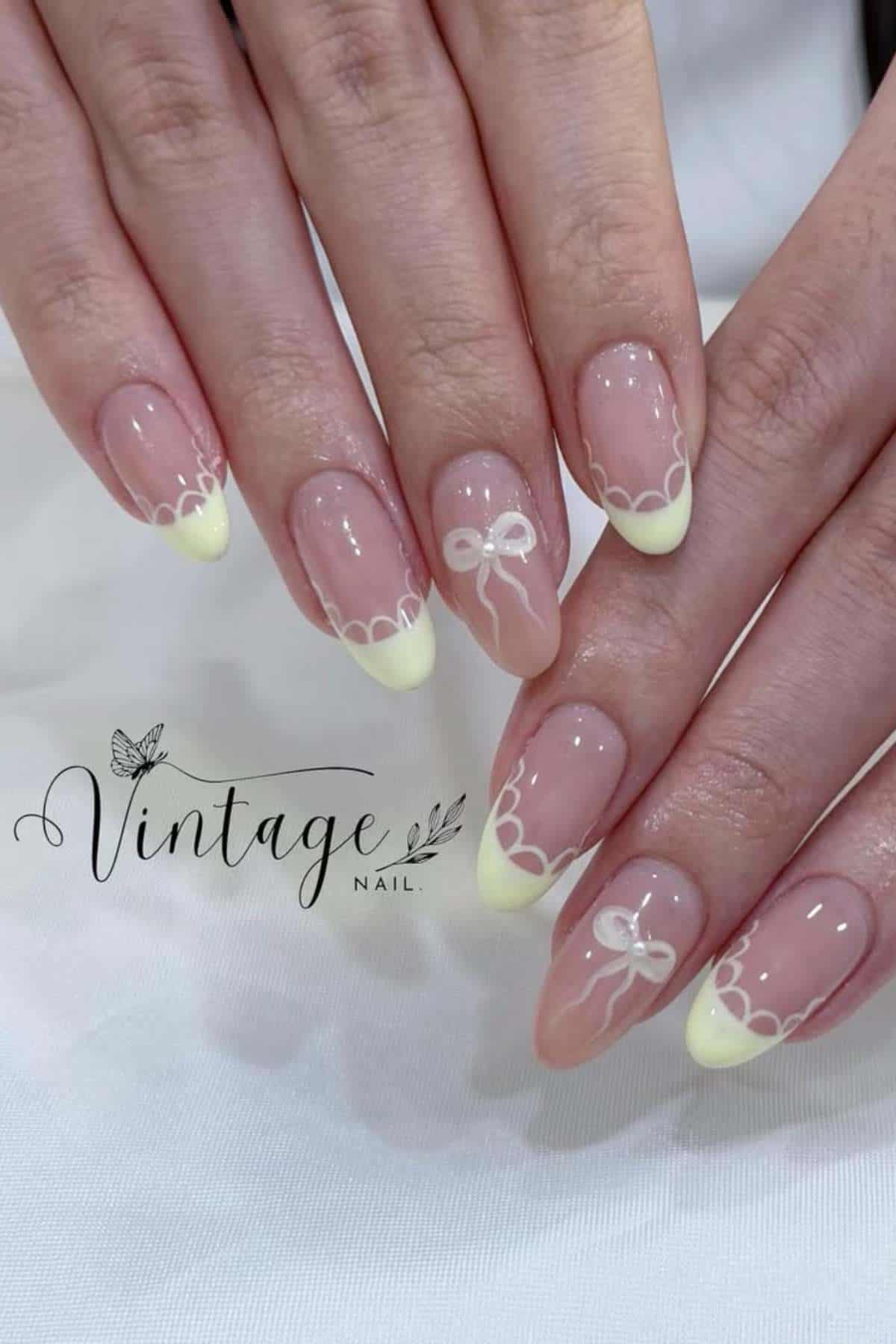 Hands with almond-shaped nails featuring pale yellow French tips, white lace-like edges, and white bow designs on a few accent nails. The "Vintage Nail" logo is visible.