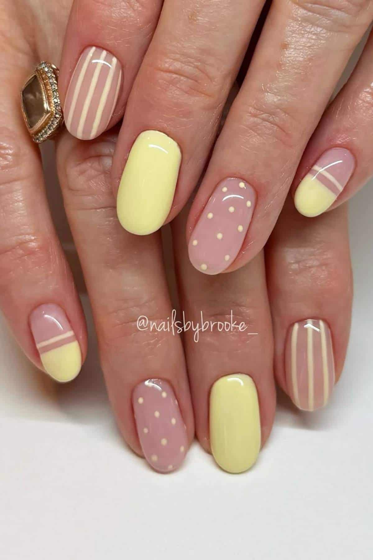 Hands with manicured nails featuring pastel yellow polish, white polka dots, and white stripes in varying designs on each nail.