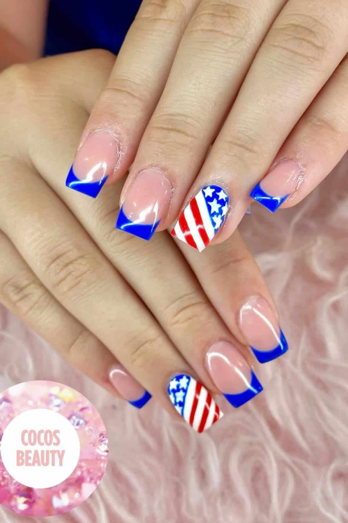 A hand with French tip nails in blue, featuring one accent nail with an American flag design of red stripes and blue stars.