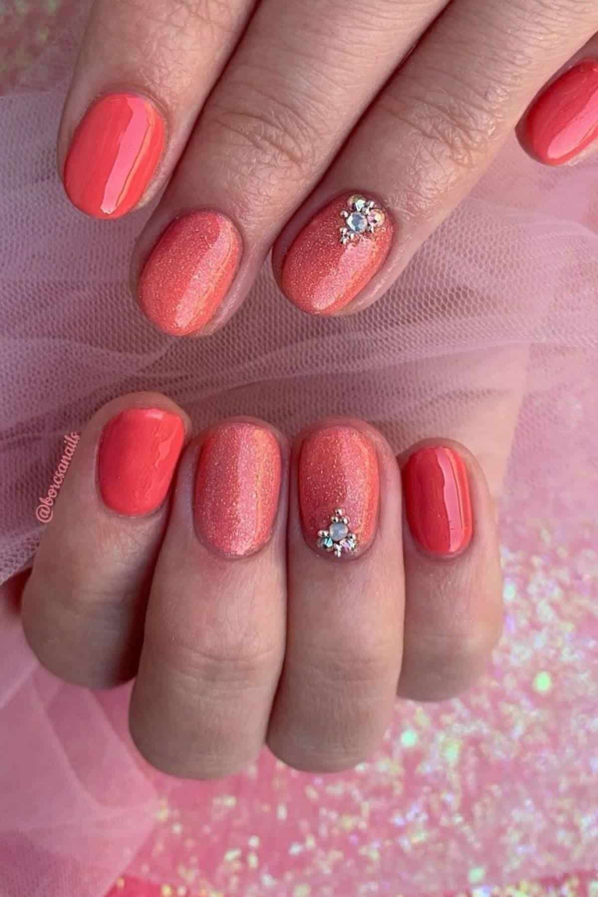 Hands with short, oval-shaped nails painted in glossy coral shades; two nails have small rhinestone flower decorations near the cuticle.