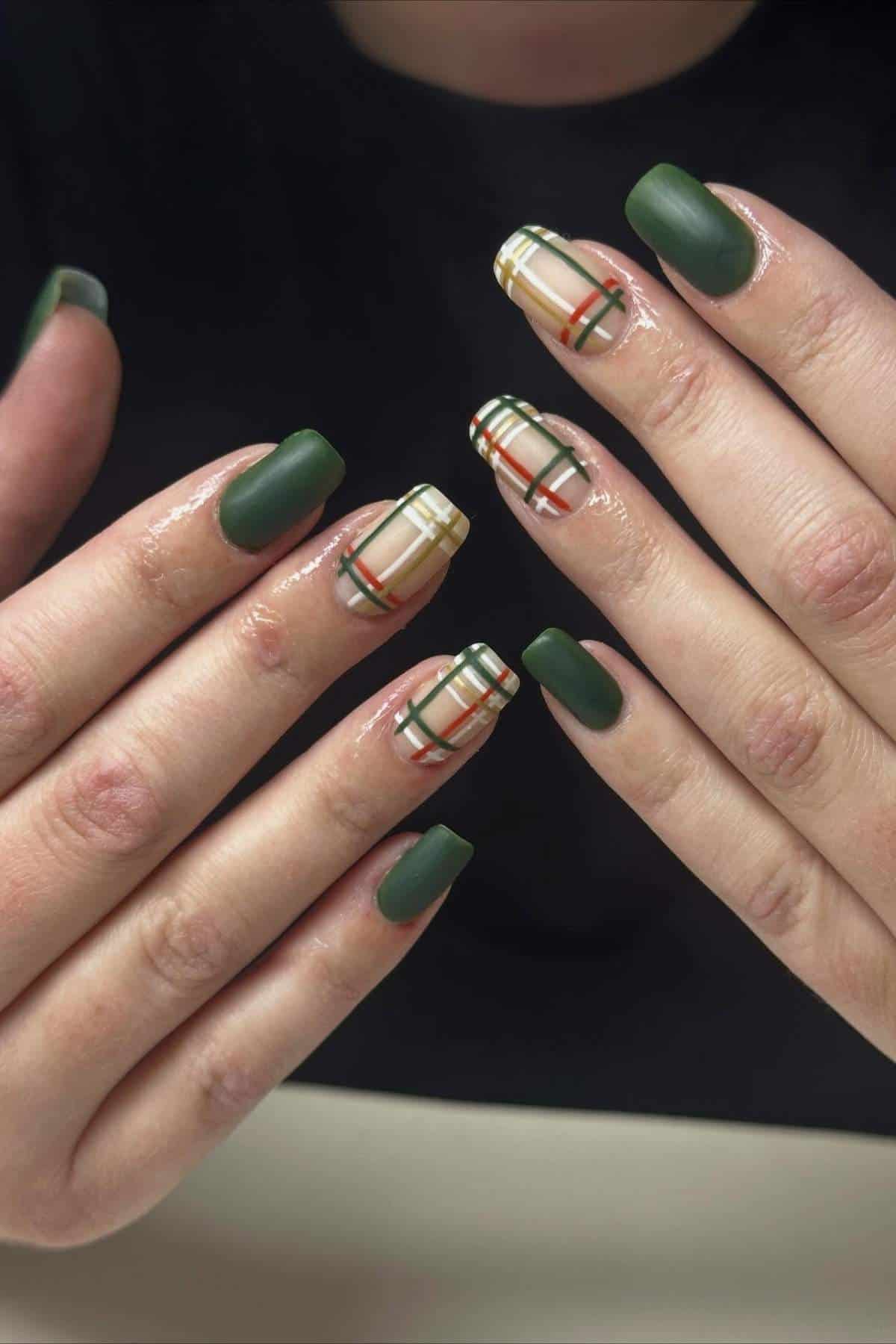 Hands with manicured nails; most nails are painted matte green, while two feature a plaid pattern with beige, white, black, and red lines.