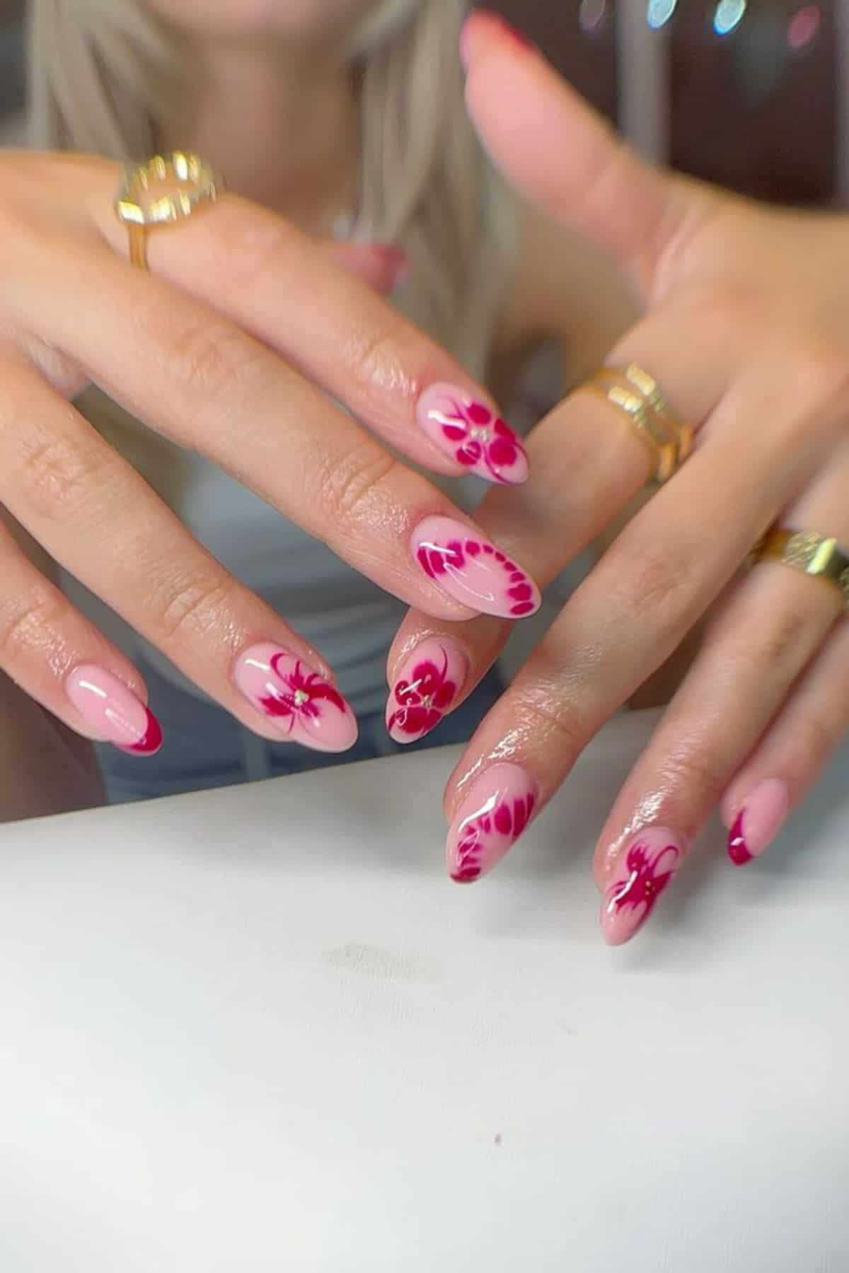 Close-up of hands with long almond-shaped nails painted light pink and decorated with red floral designs; person is wearing several gold rings.