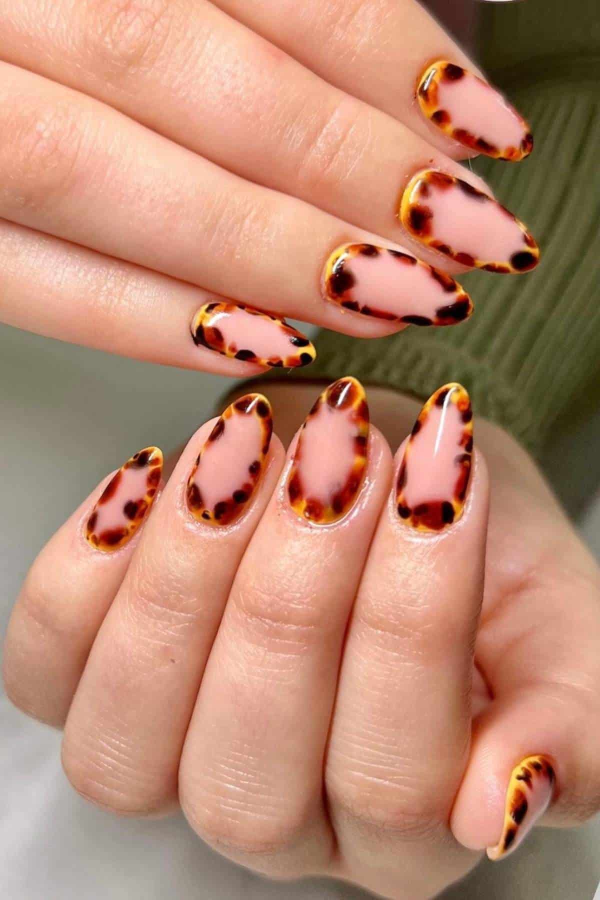 Close-up of hands with almond-shaped nails featuring a pink base and tortoiseshell-patterned tips.