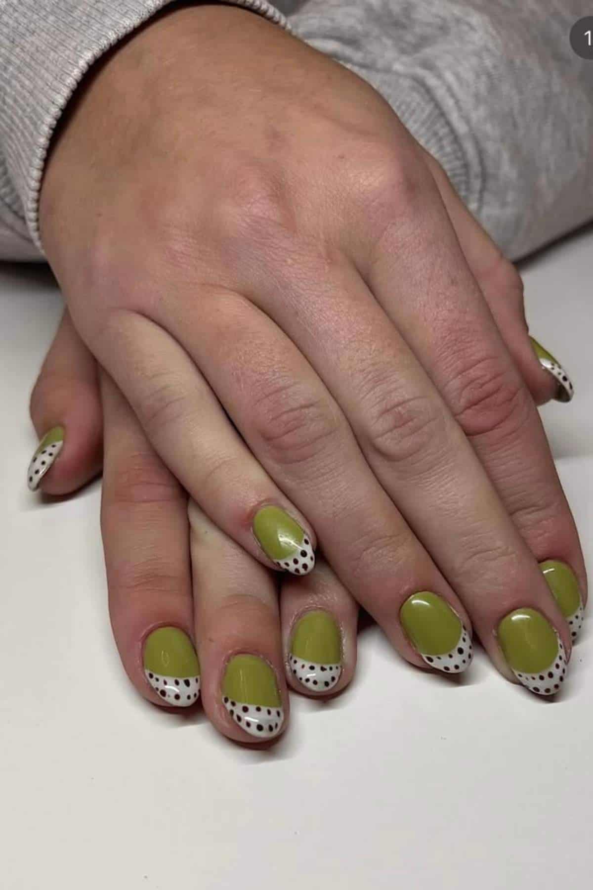 Hands with green nail polish and white tips decorated with black polka dots, resting on a white surface.
