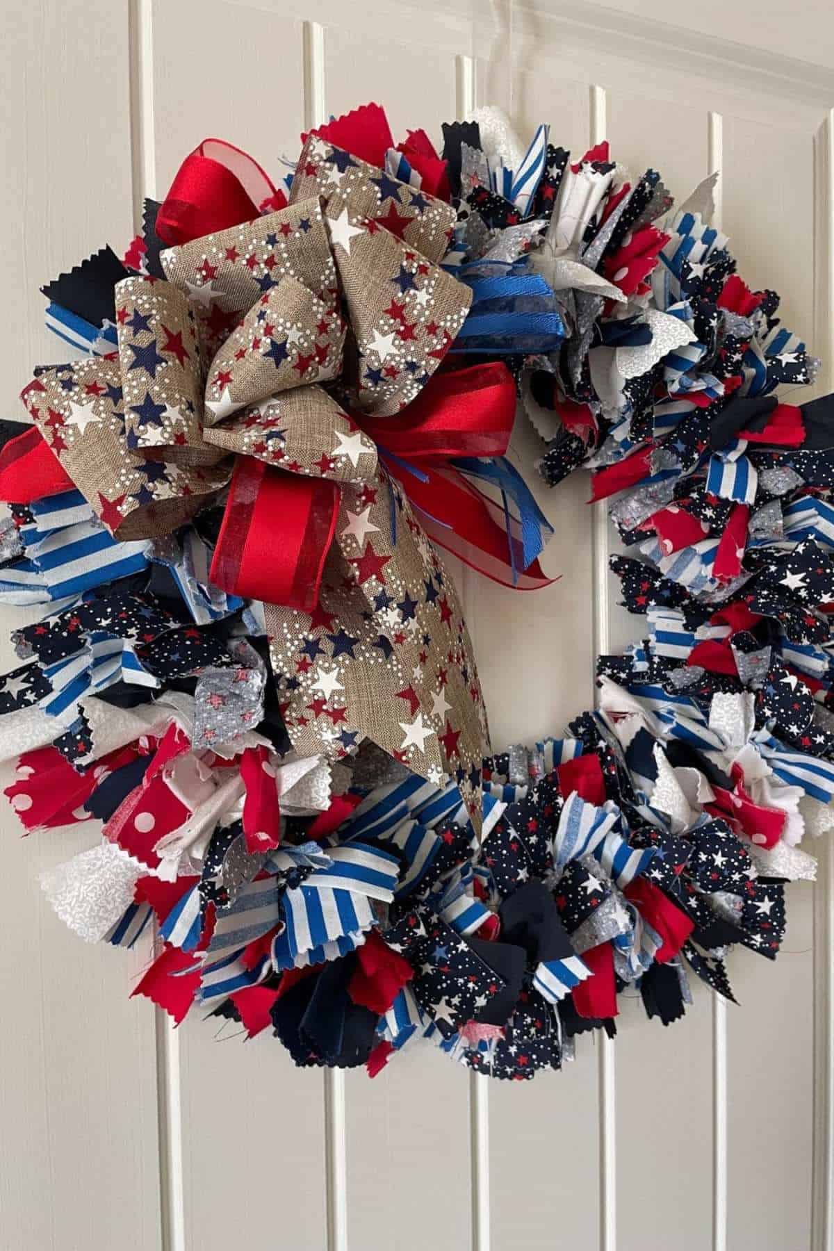 A wreath made of red, white, blue, and silver fabric strips with star patterns, accented by a large bow, hangs on a white paneled door.